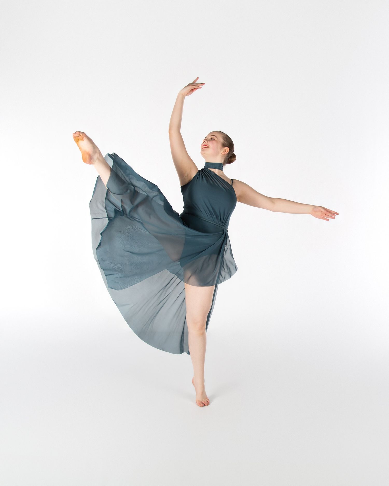 Dancer in grey flowing dress performs an elegant arabesque position against white backdrop.
