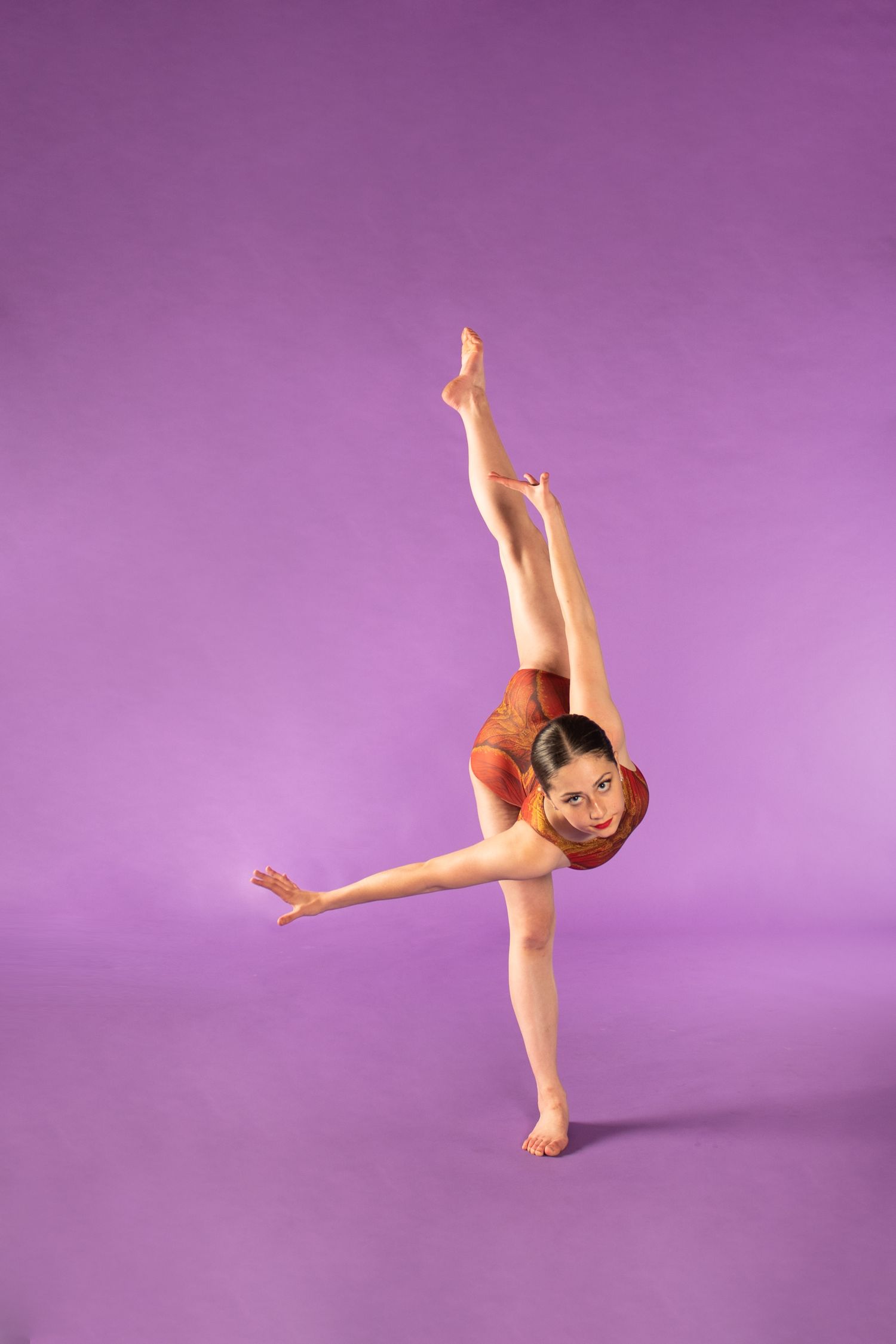 Young gymnast in red leotard performs graceful balancing pose against purple background.