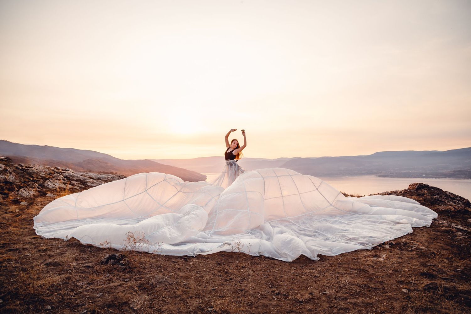 A bride in a flowing white dress with a dramatic circular train poses against a mountain landscape at sunset.