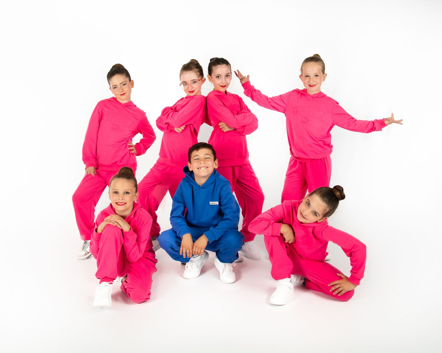 Dance group in bright pink tracksuits and one blue tracksuit poses together against white backdrop.