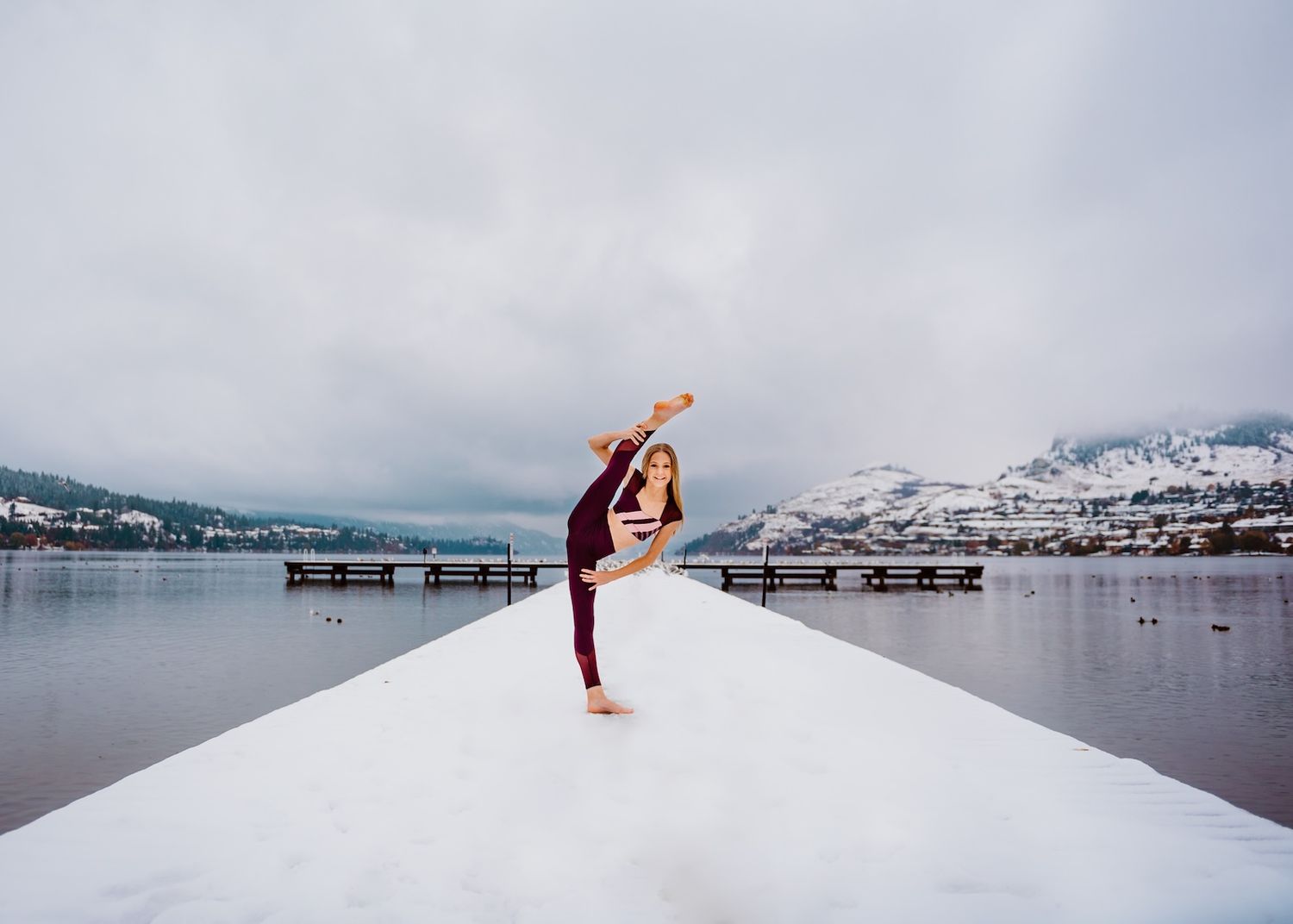 Dancer performing graceful pose on snowy dock with mountains and lake in winter landscape.