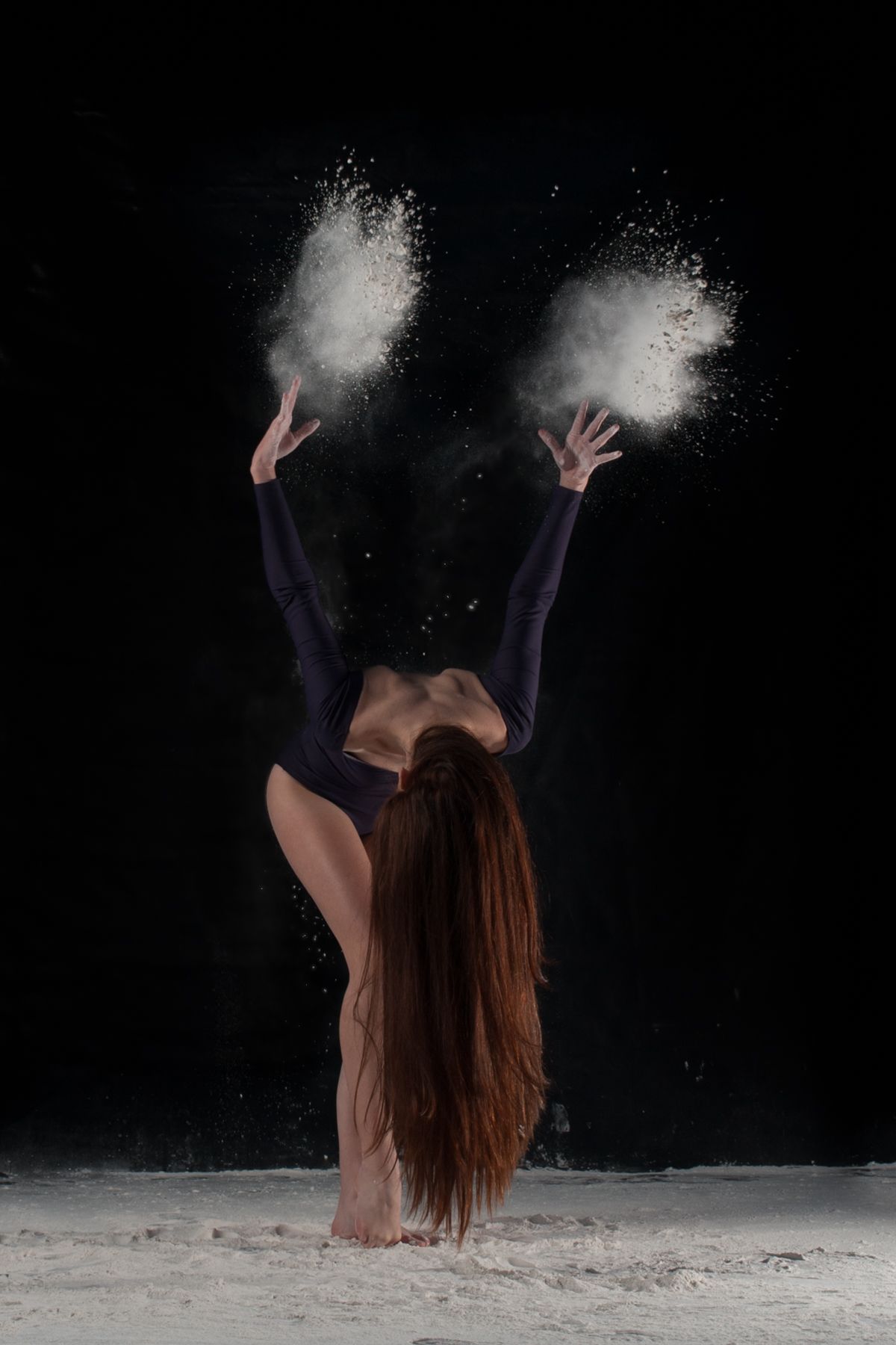 Dancer in black bodysuit creates dynamic pose with flour powder effects against dark background.