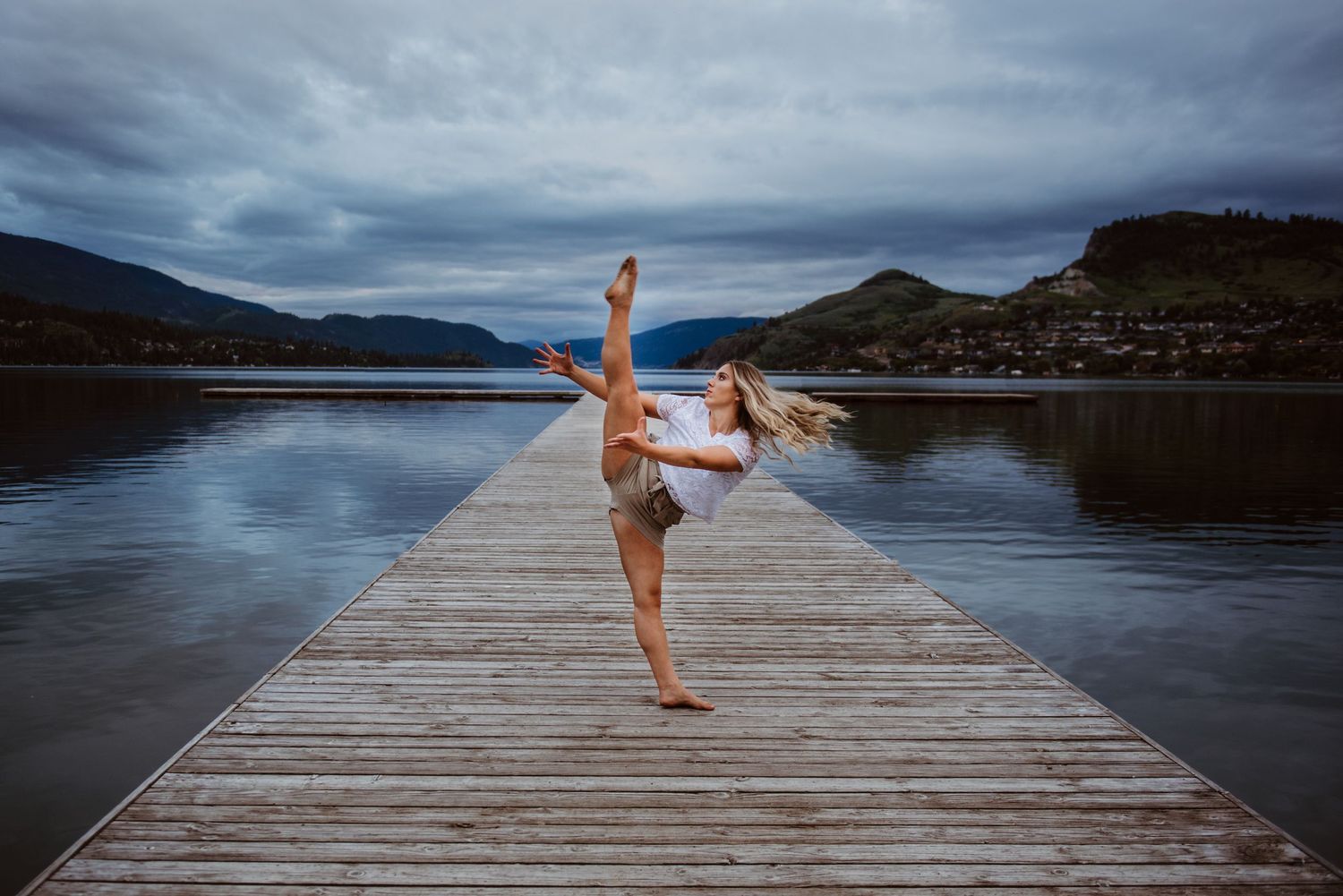 Dancer performs graceful pose on wooden pier overlooking mountain lake under dramatic cloudy sky.