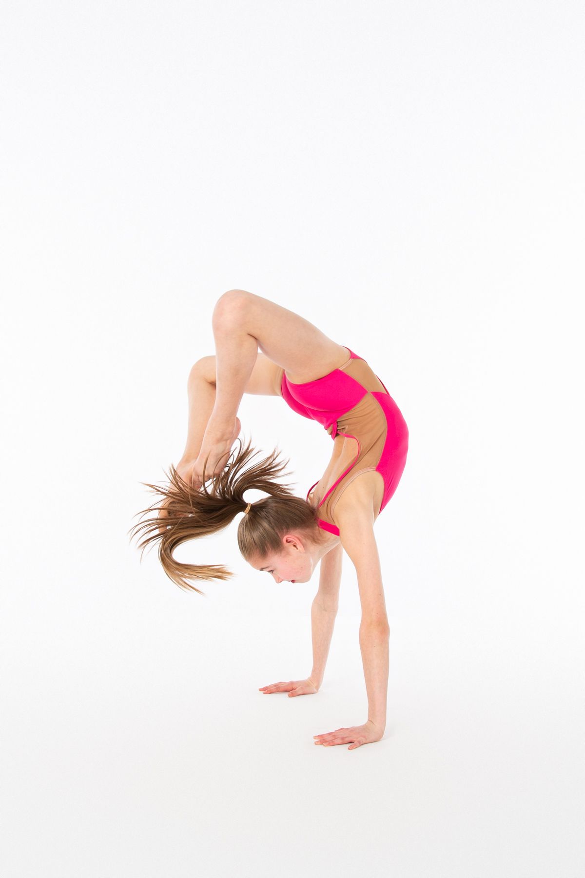 Young gymnast in pink leotard performing a backbend handstand on white background.