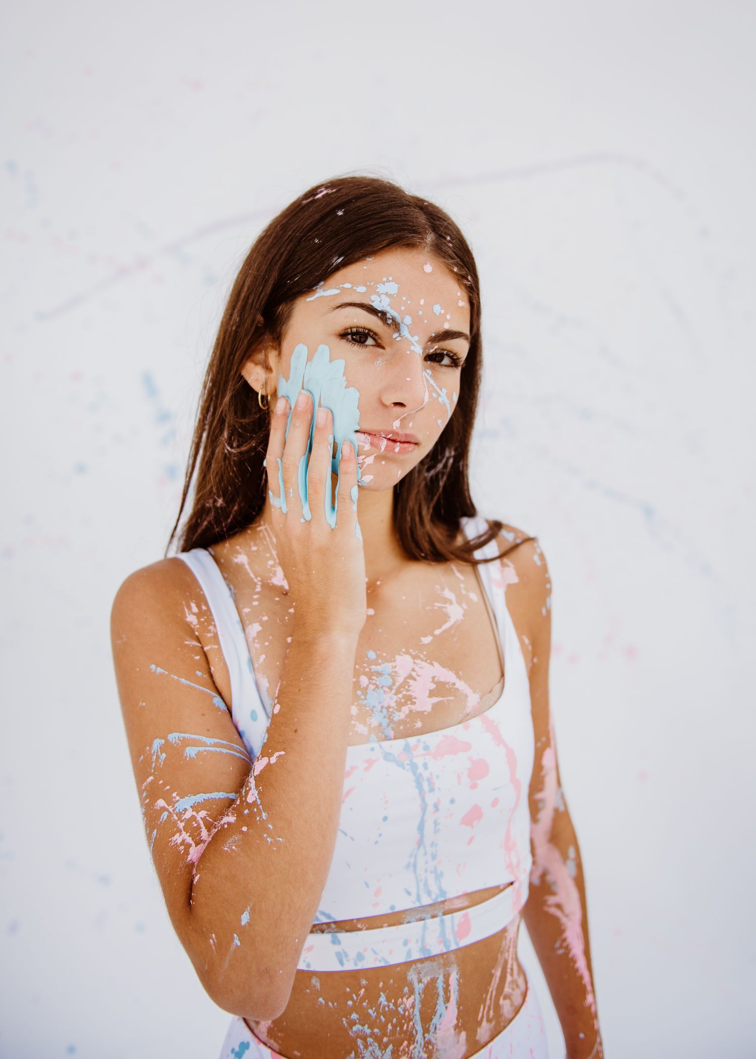 Person in white outfit poses artistically with paint splattered on skin against light colored background.
