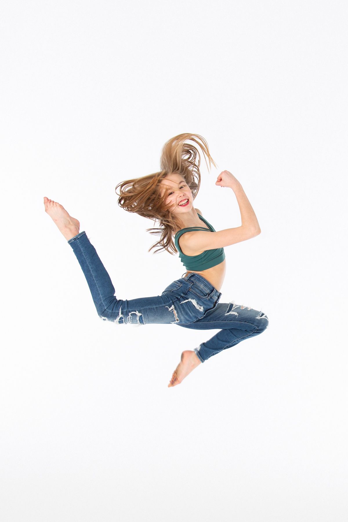Energetic dancer in blue jeans and green top leaps joyfully against white background with hair flowing in motion.