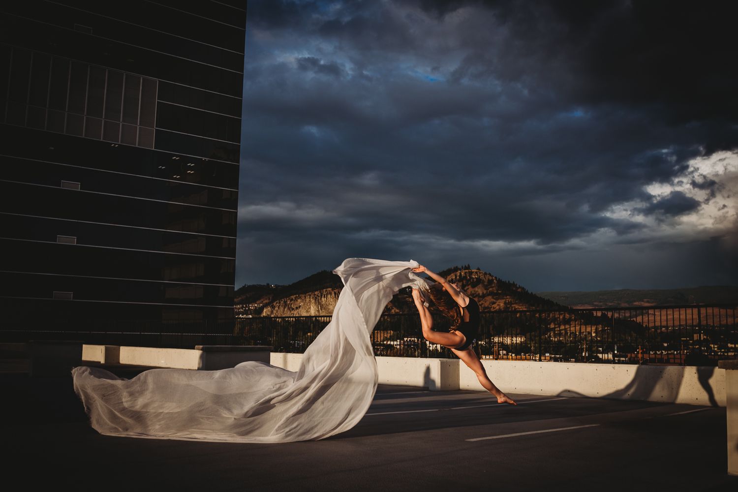 A dramatic wedding photo with a flowing white veil billowing against dark storm clouds and mountain backdrop at sunset.