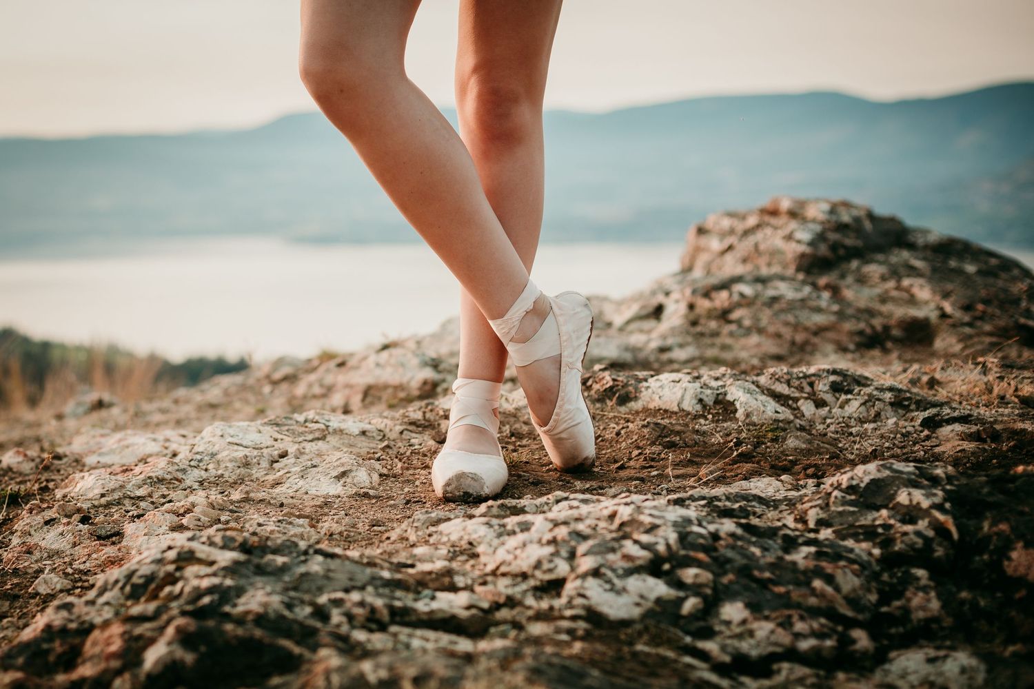 Legs wearing white sneakers stand on rocky cliff edge overlooking misty blue mountains and sea at sunset.