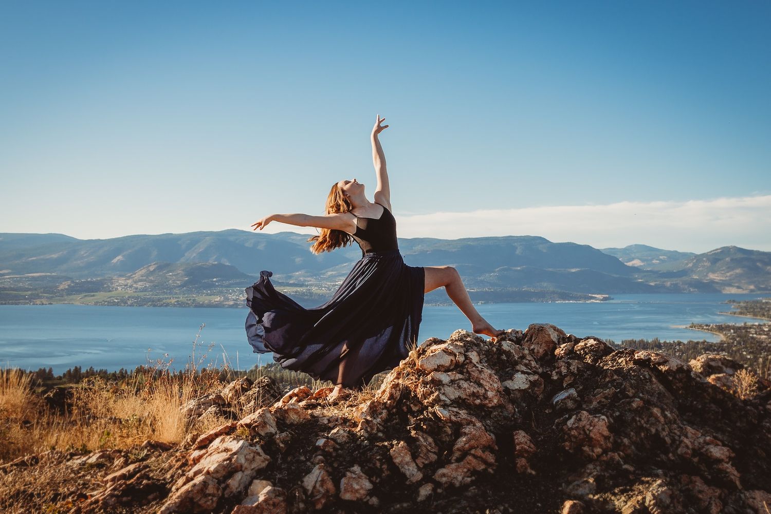 A dancer in a flowing black dress leaps gracefully on rocky terrain overlooking a lake and mountain landscape.
