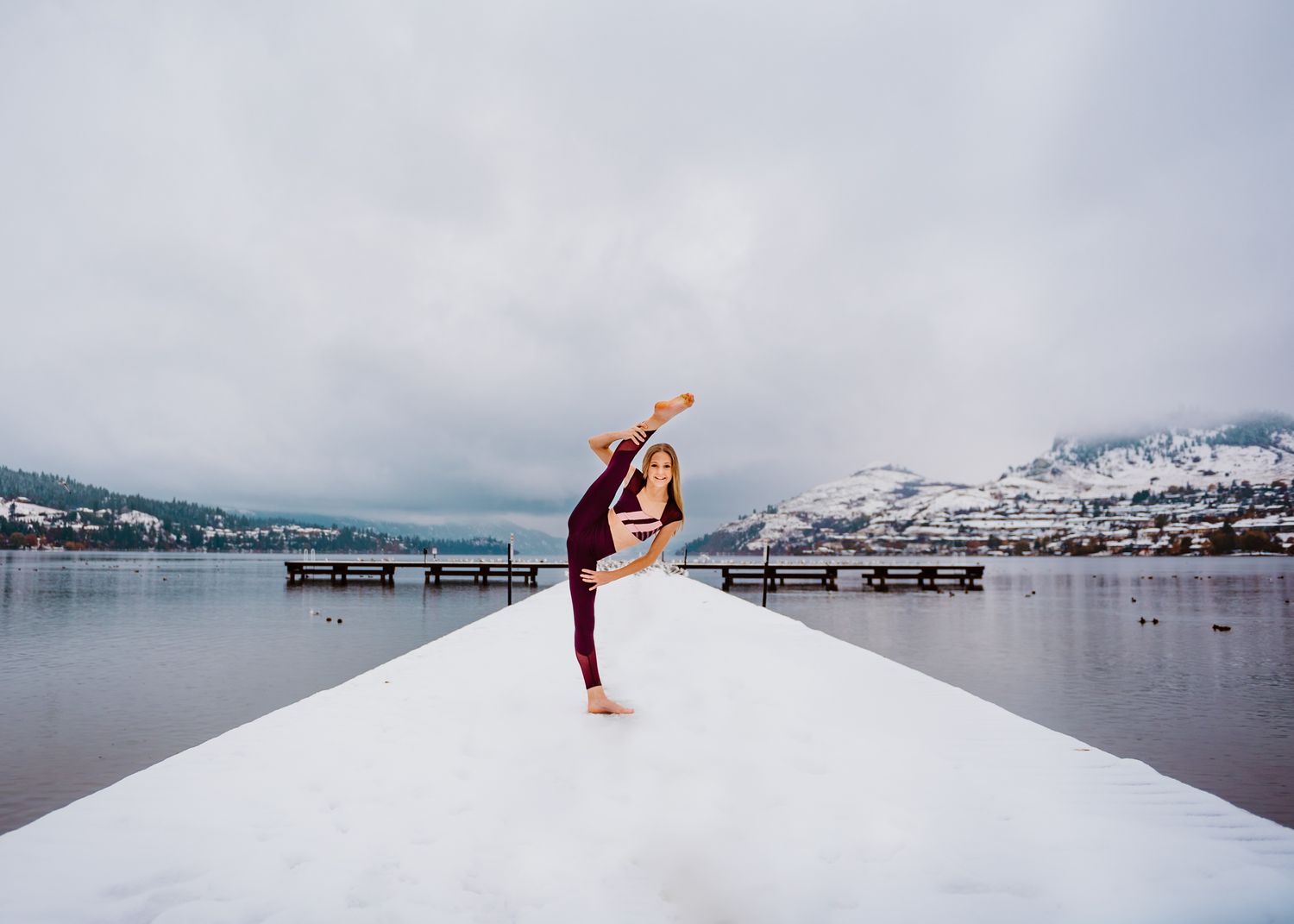 A person performs a graceful dance pose on snowy ground against a scenic mountain lake landscape.