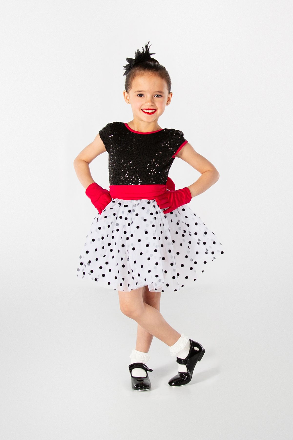 Dance performer wearing black sequin top, polka dot tulle skirt, red gloves and tap shoes poses against white background.