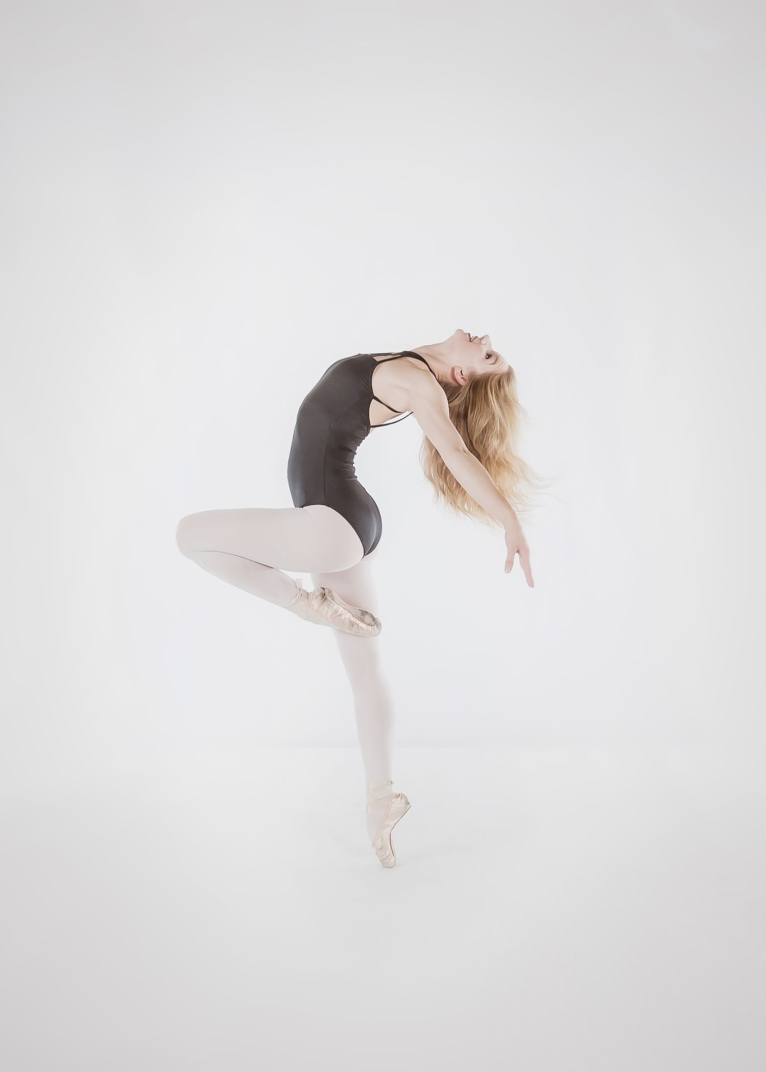 A ballet dancer in a black leotard and pink tights performs a graceful pose while balancing en pointe against a white backdrop.