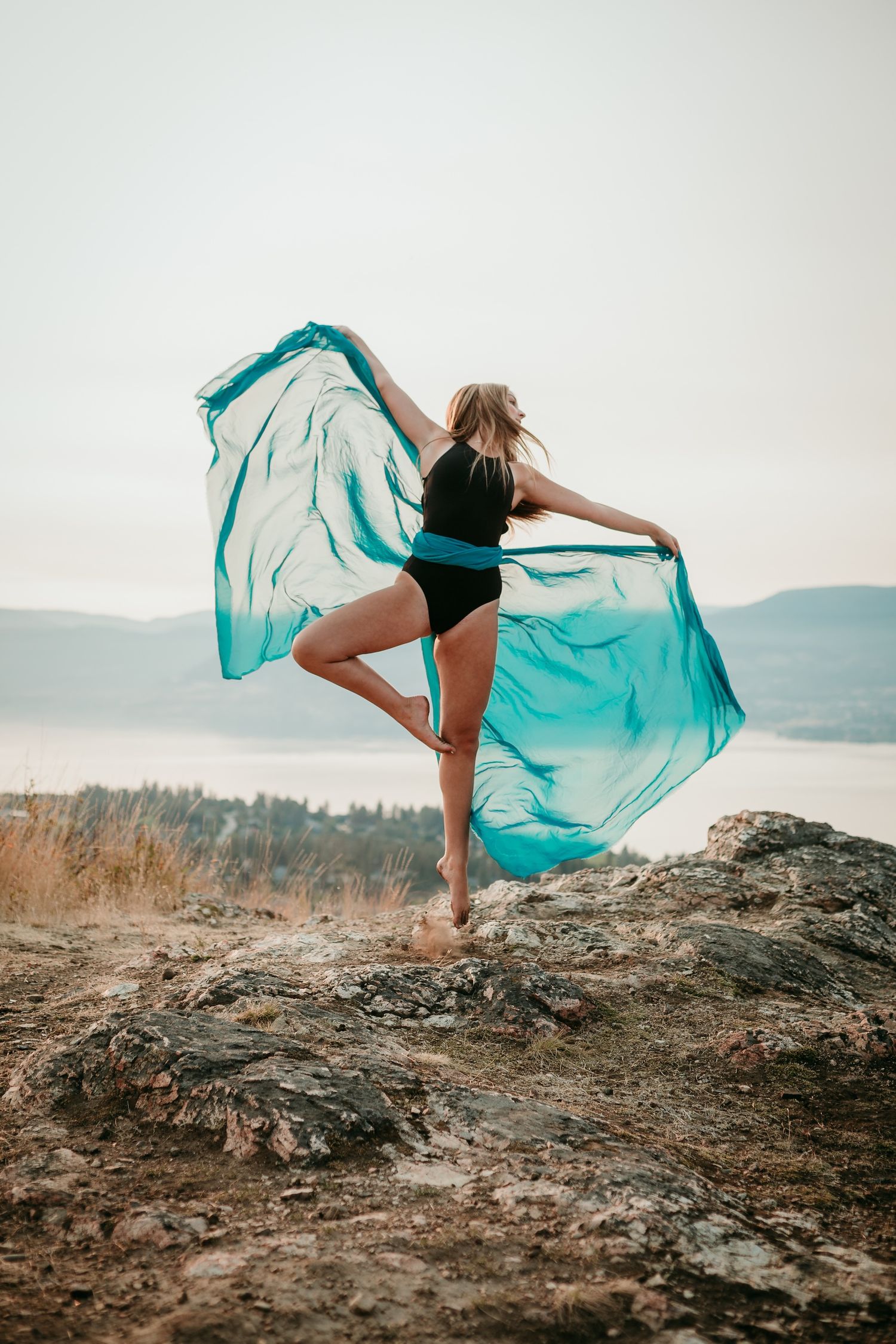 Dancer performs on rocky cliff with billowing turquoise fabric against mountain vista.