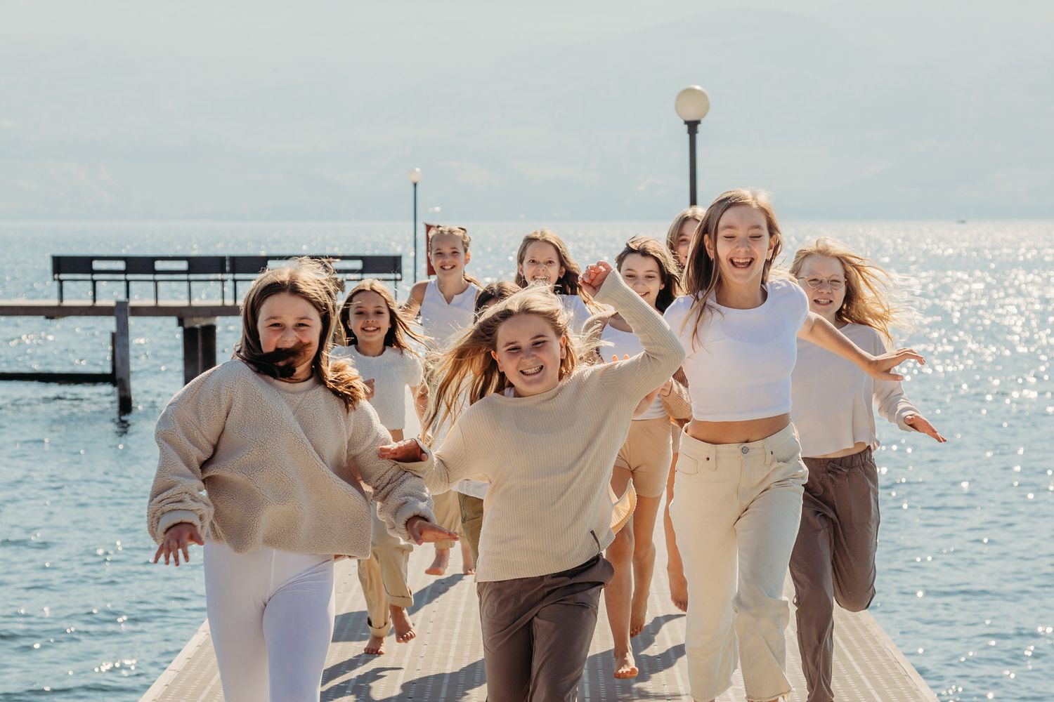 Group in neutral-toned clothing poses joyfully on a pier with sparkling water and sunlight behind them.