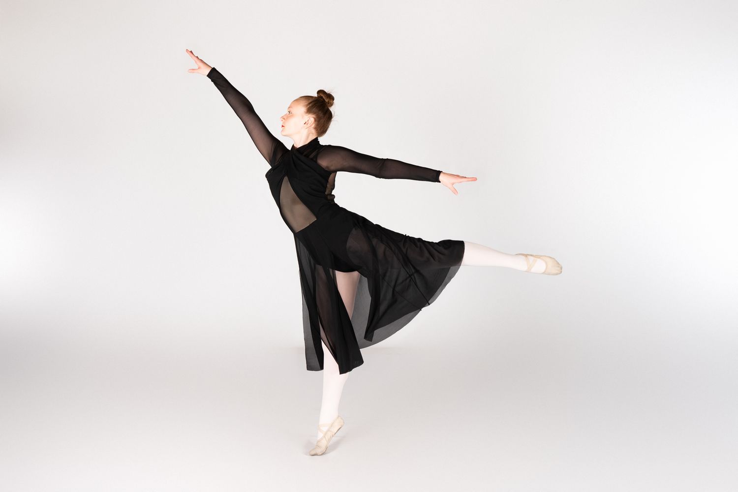 Ballet dancer in flowing black dress performs graceful pose en pointe against white backdrop.