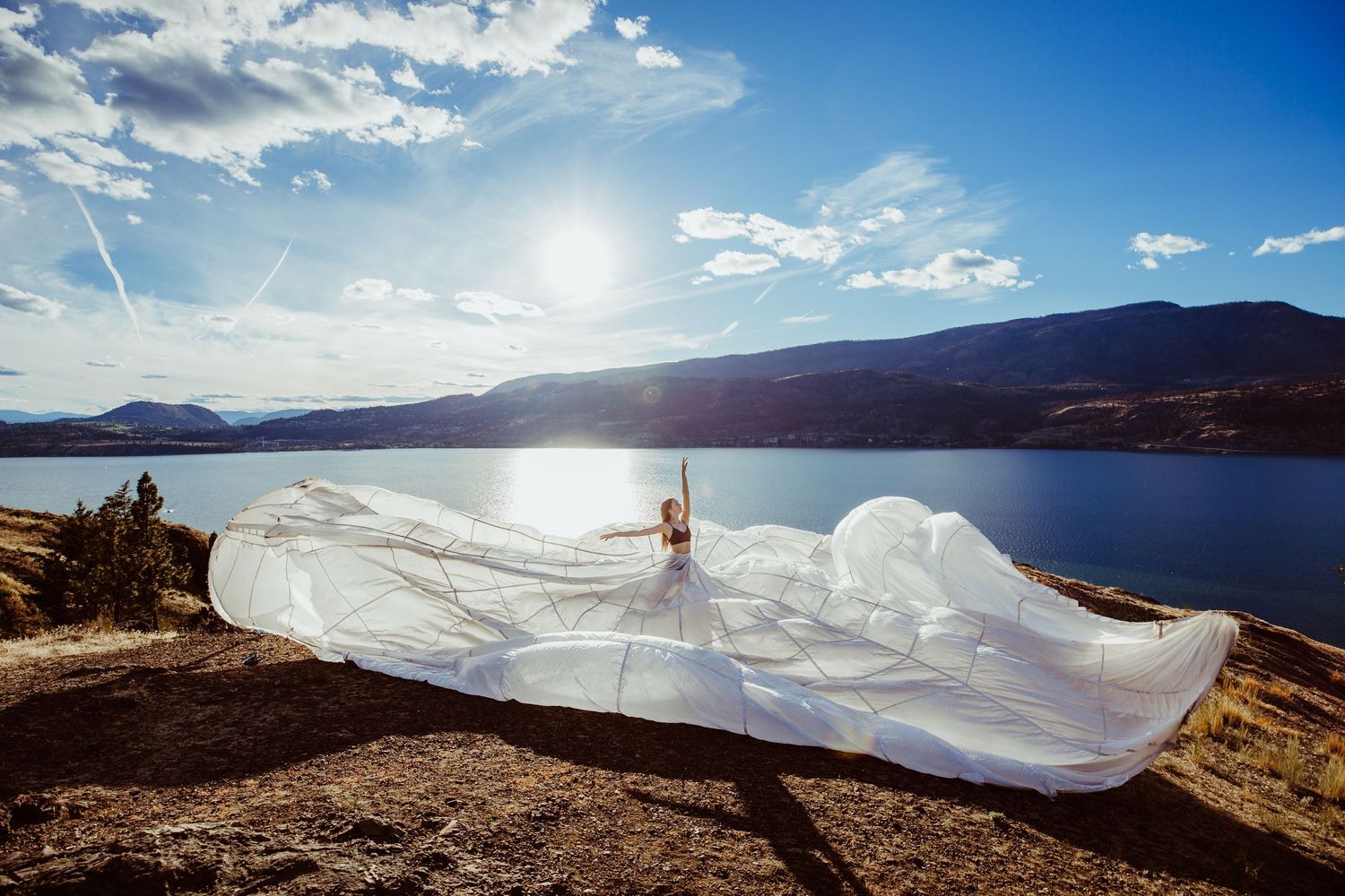 Flowing white fabric billows dramatically against a sunlit mountain lake landscape with blue skies.