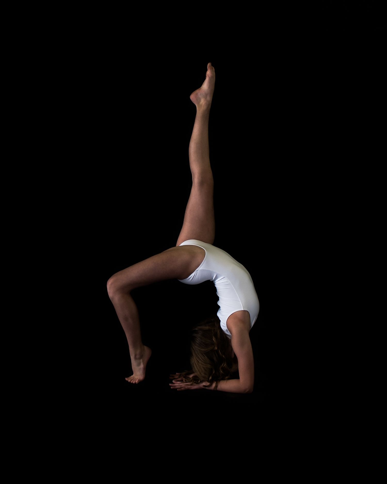 Gymnast in white leotard performs graceful backbend against black background in dramatic artistic pose.