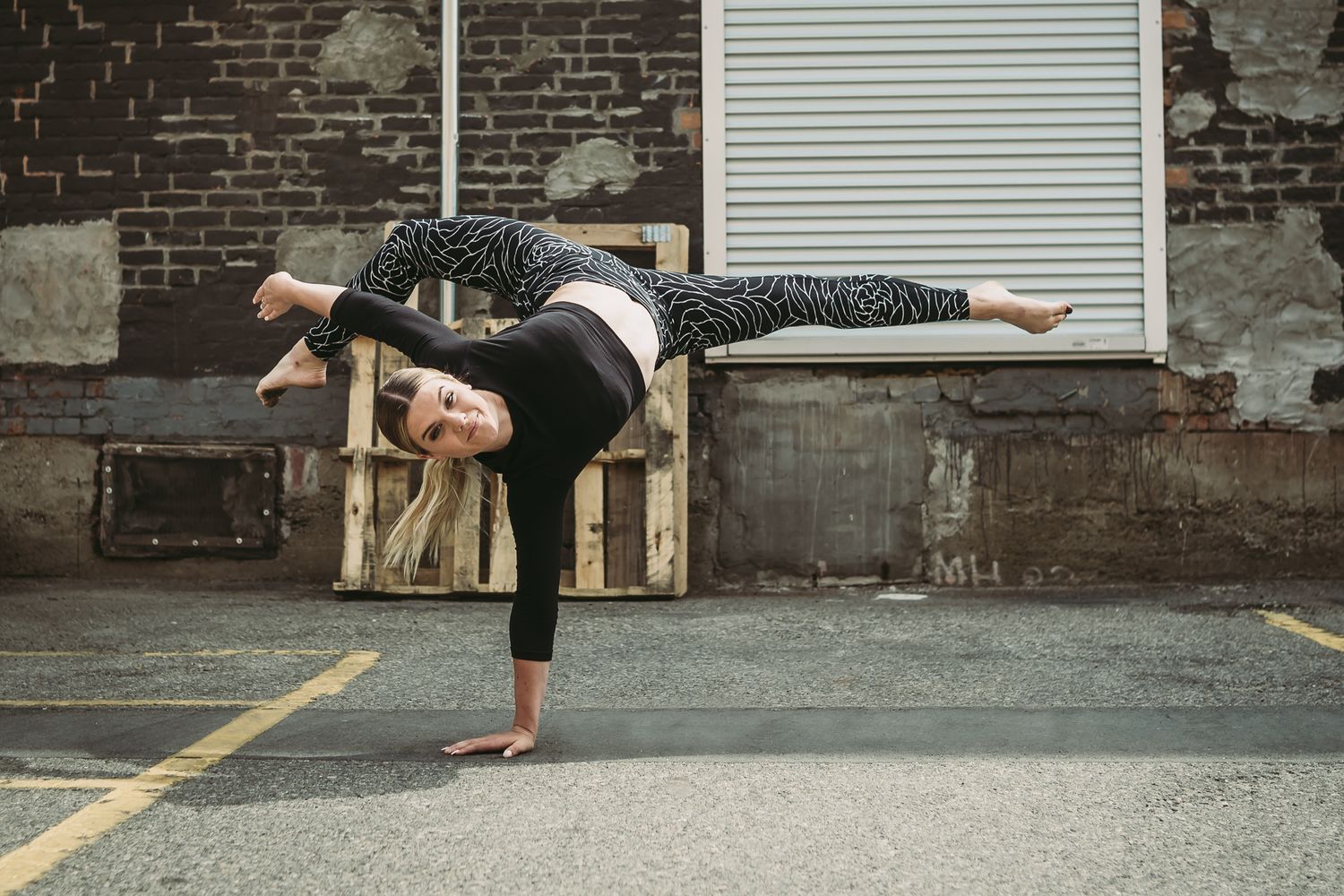 Dancer in black athletic wear performs a graceful standing split pose against an urban brick wall backdrop.