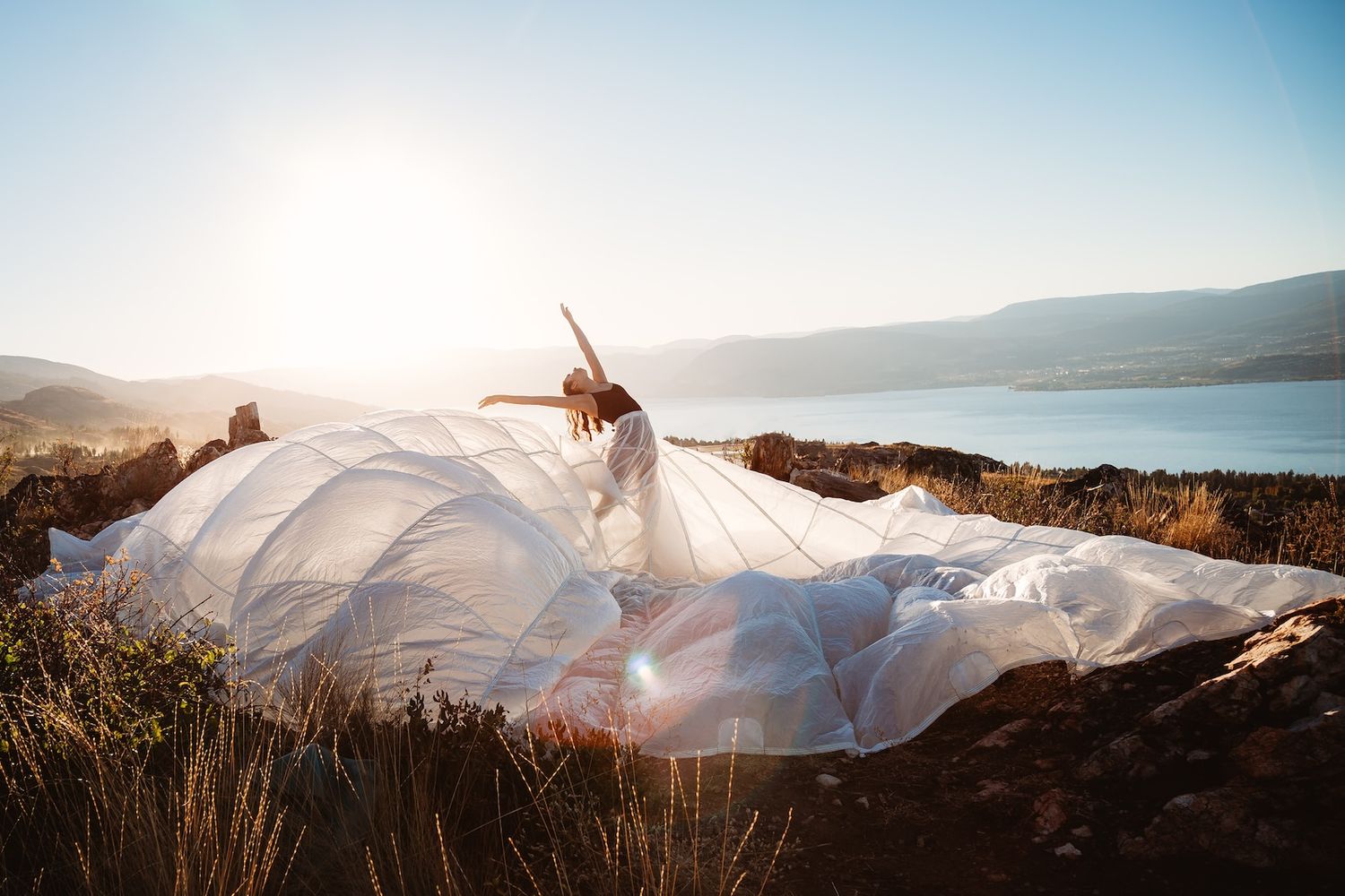 A flowing white dress billows dramatically against a scenic mountain landscape at sunset.