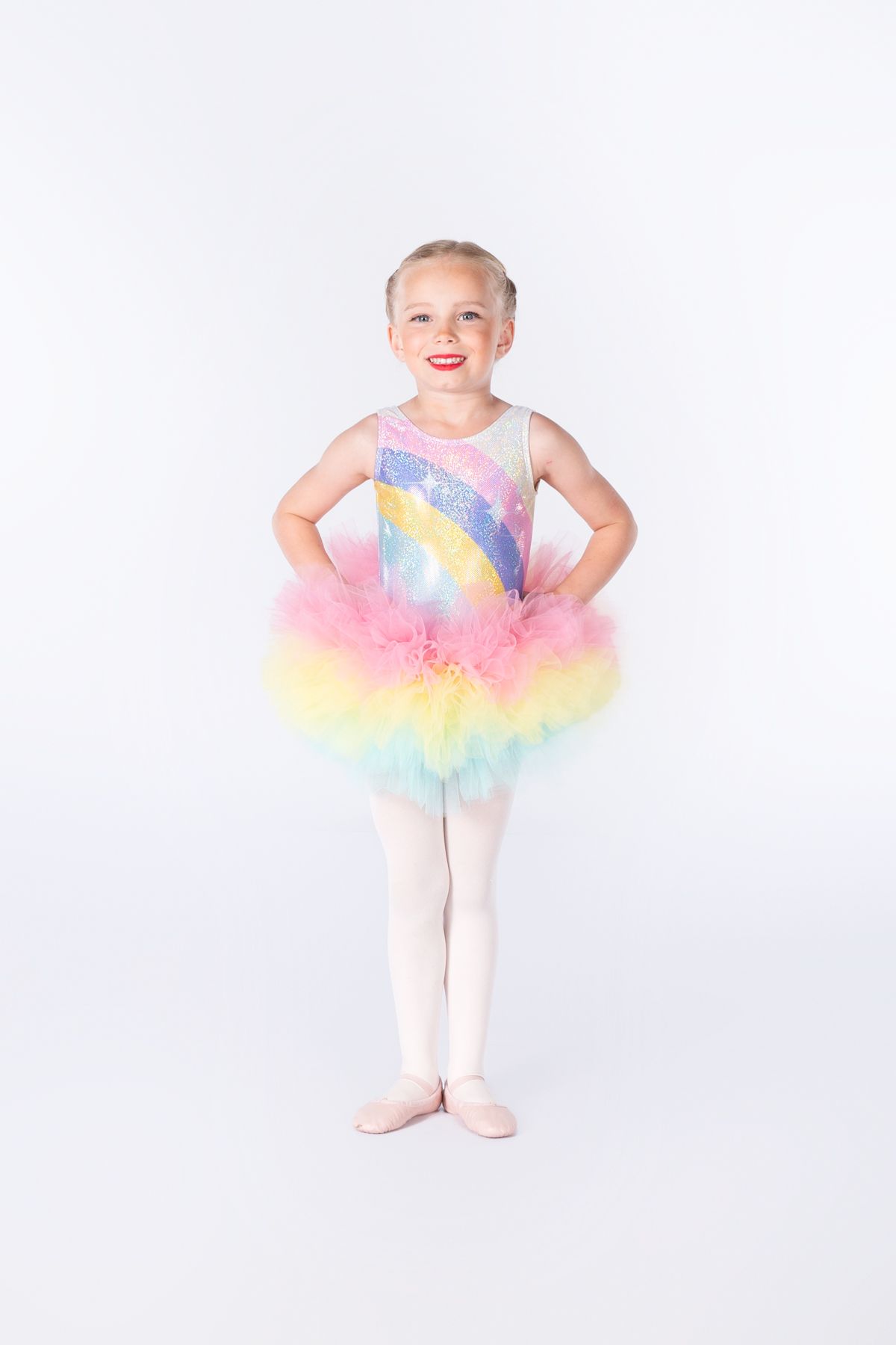 Young dancer strikes a ballet pose wearing a pastel rainbow tutu and leotard against a white background.