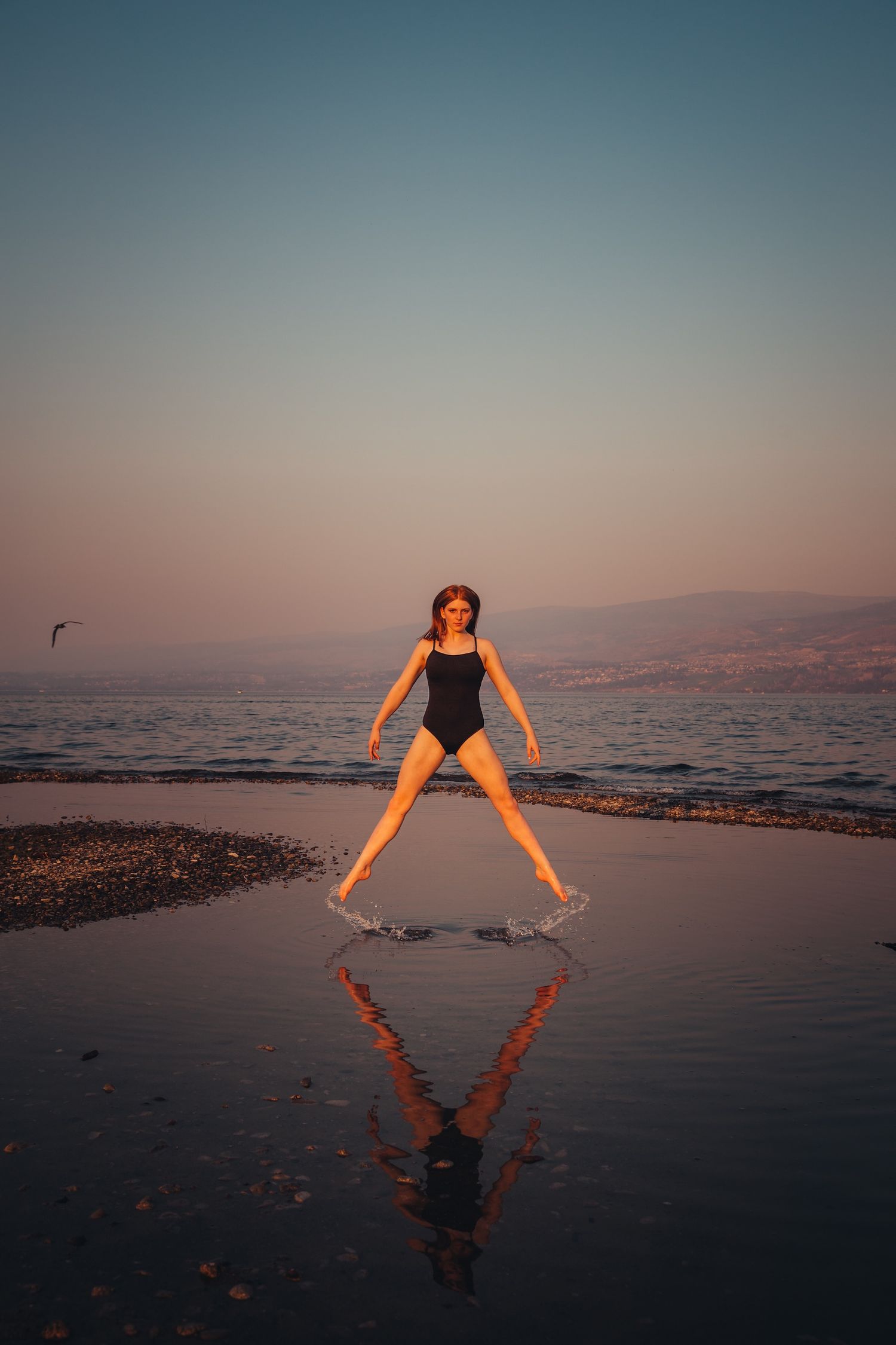 A dancer leaps on a beach at sunset creating a reflection in the wet sand as a bird flies in the pink and blue sky.