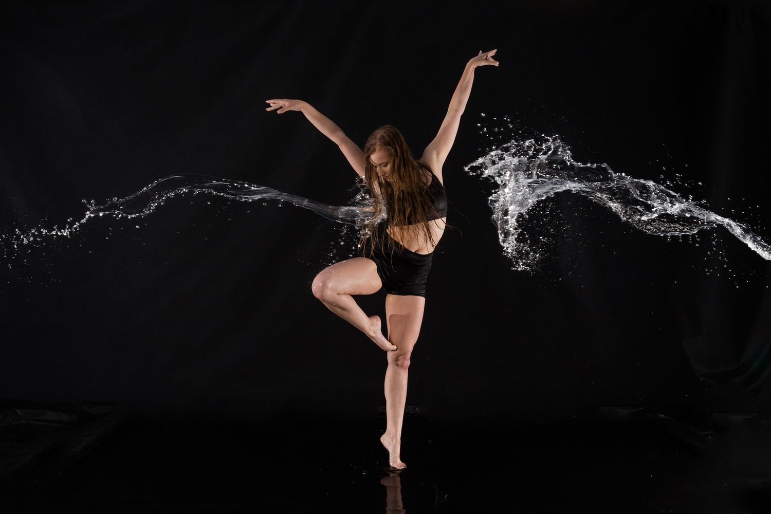 Dancer in black performing graceful pose with water splashing dramatically against dark background.