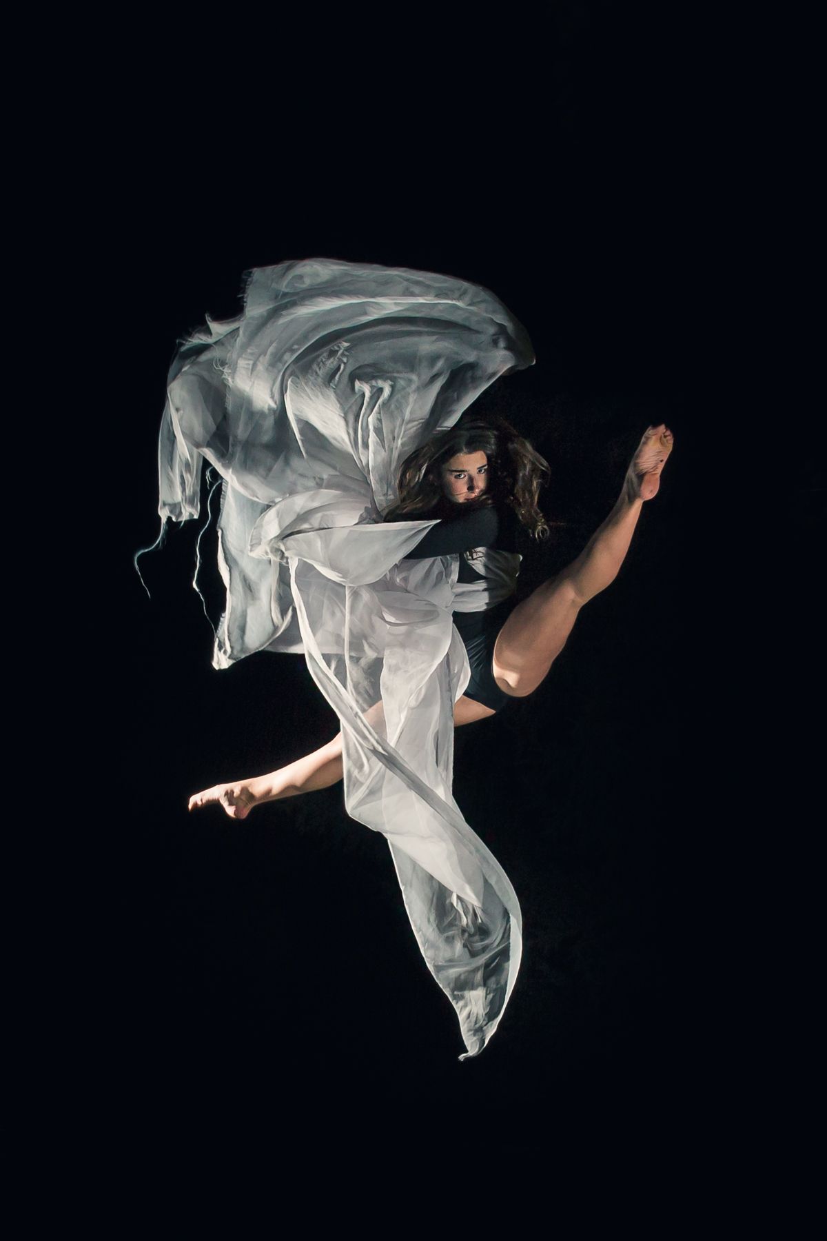 Dancer in flowing white fabric leaps gracefully against a dark background in dramatic artistic performance shot.