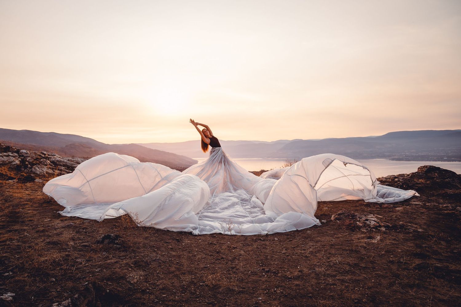 A flowing white wedding dress billows dramatically against a dusty mountainous landscape at sunset.