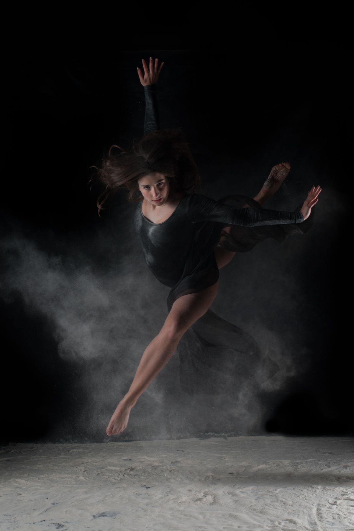 Dynamic dancer leaping through smoky air in black leotard against dark background.