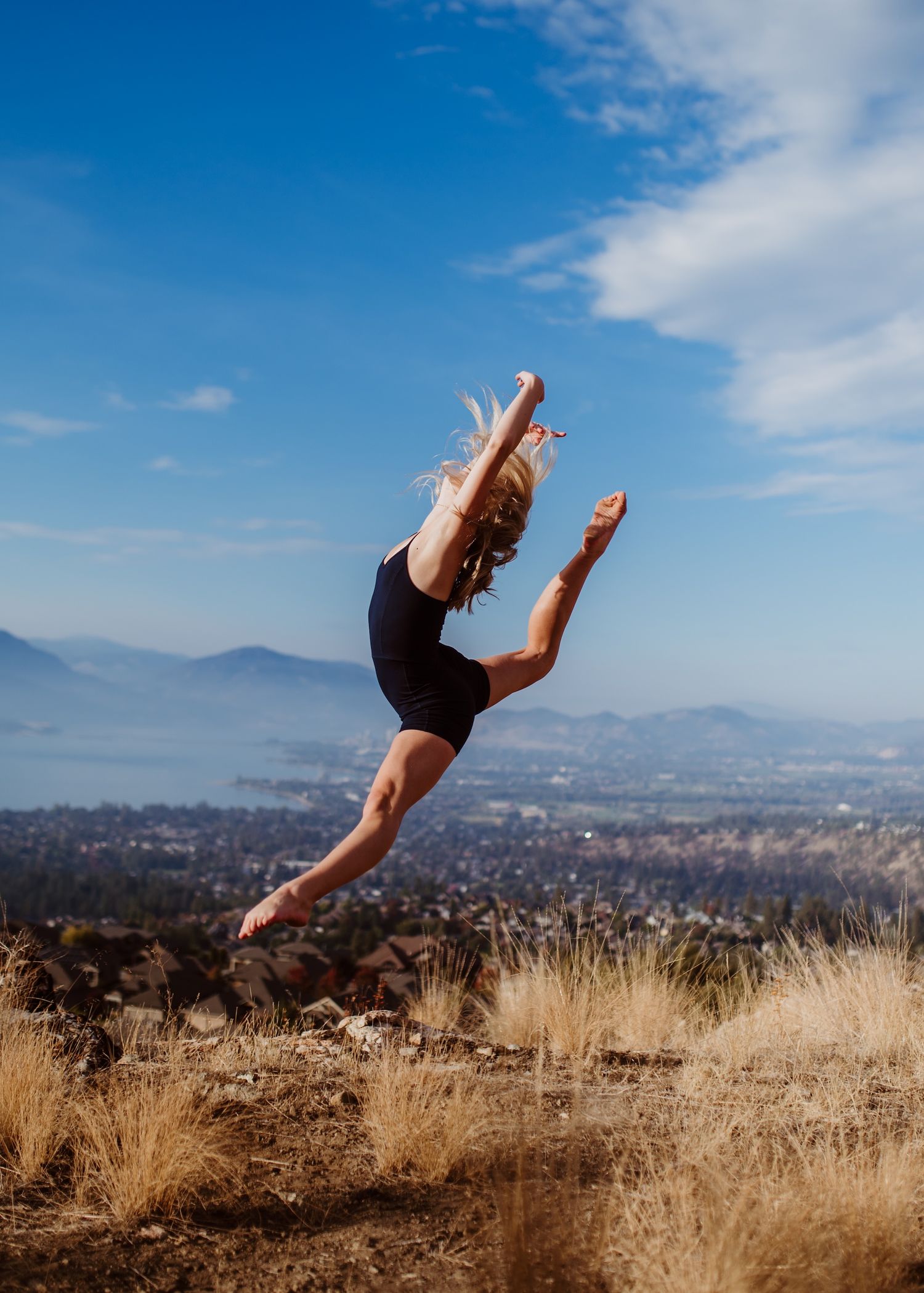 Dancer performs a dramatic leap against a mountain landscape backdrop with blue sky and dry grass.