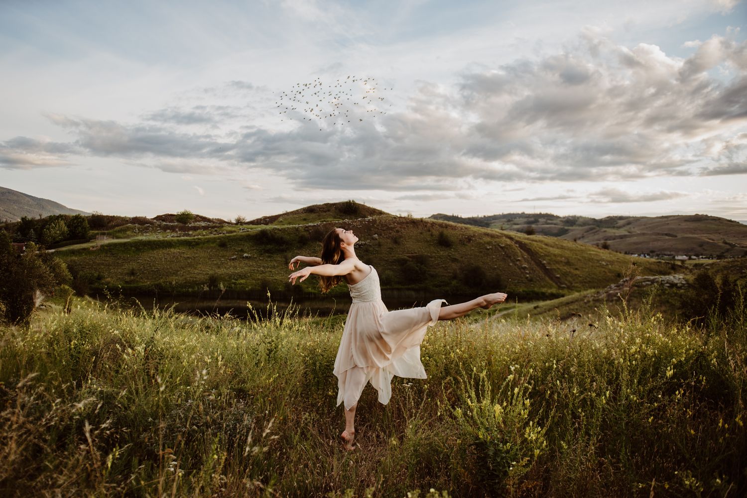 A dancer in white dress performs gracefully in a scenic meadow with rolling hills and cloudy sky at sunset.