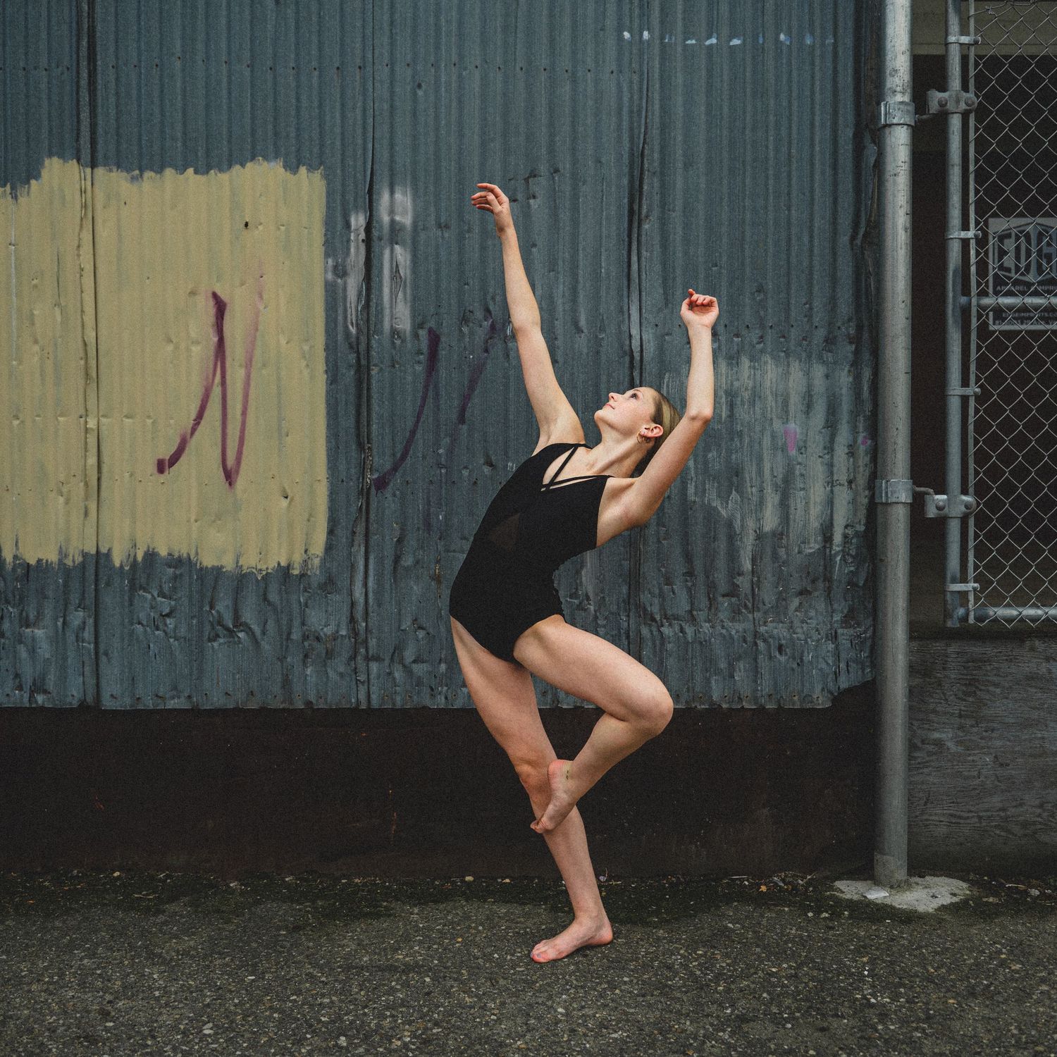 A dancer in black leotard performs an expressive pose against an industrial wall background.