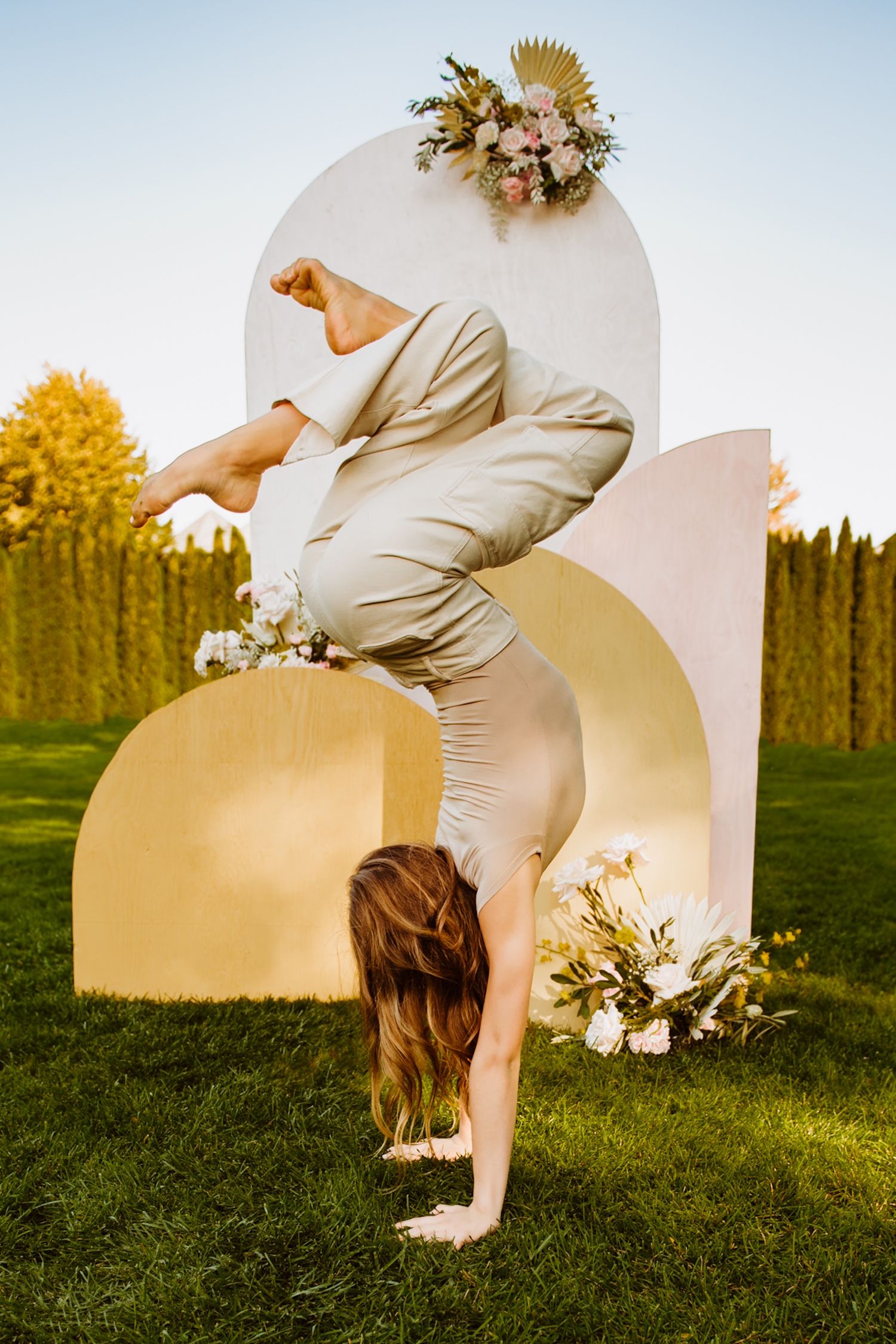 Flexible dancer performs handstand in front of curved golden backdrop with floral arrangements on green lawn at sunset.
