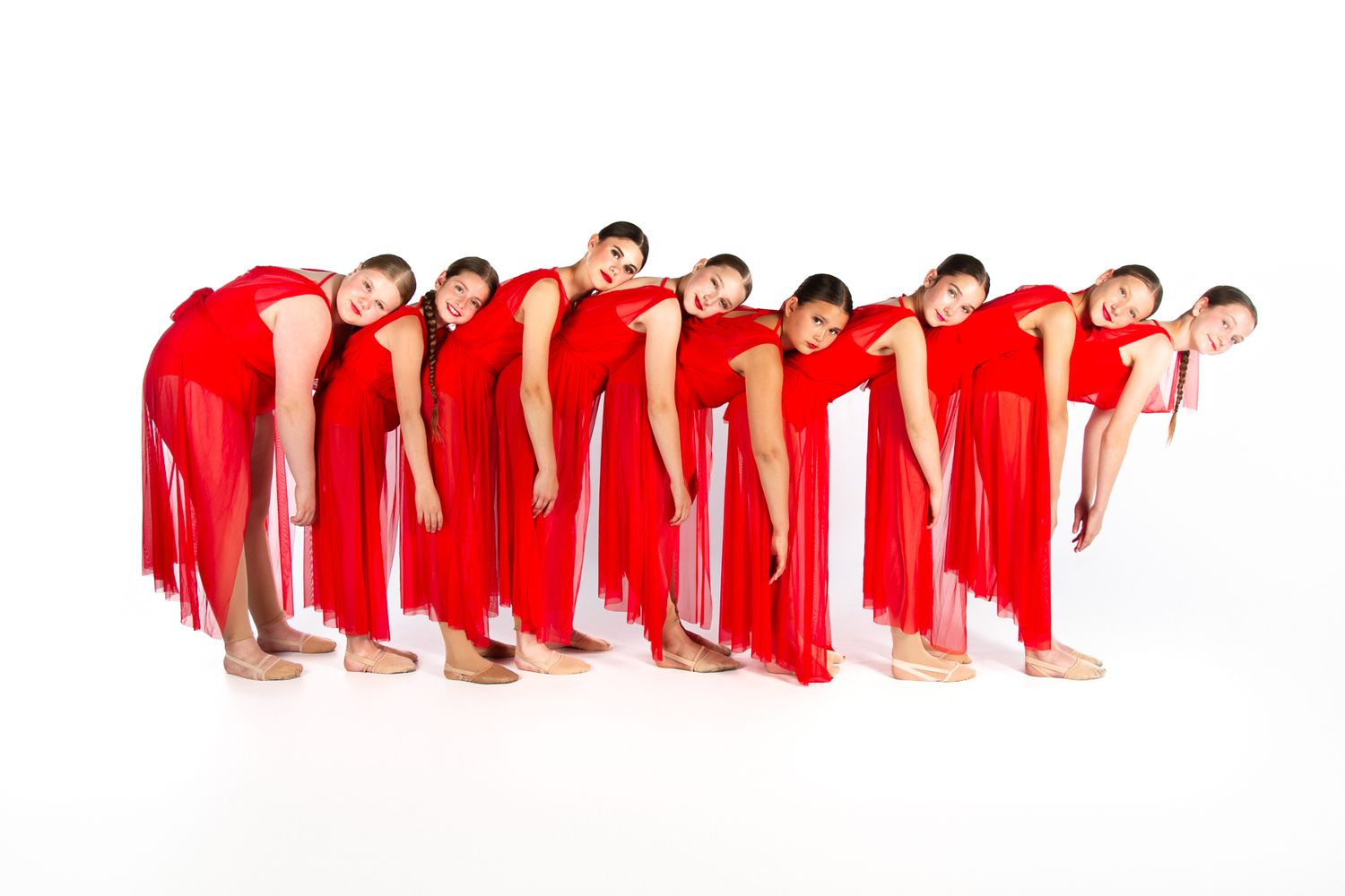 Line of dancers in bright red dresses creating synchronized leaning formation against white backdrop.