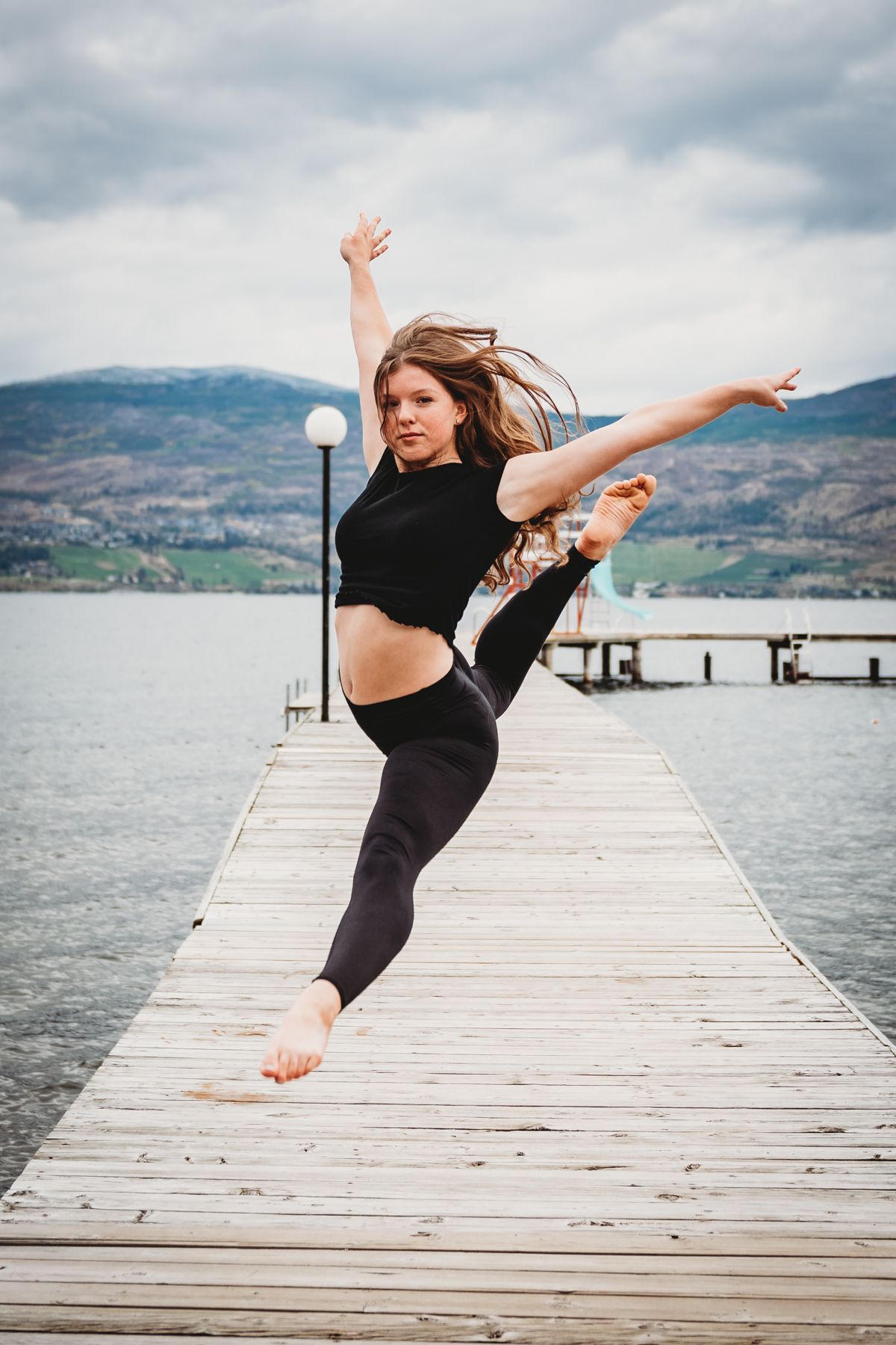 A dancer in black athletic wear performs a dramatic pose on a wooden dock overlooking a scenic mountain lake.