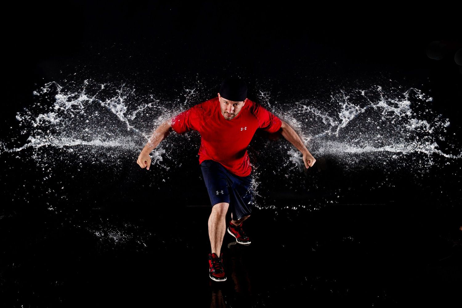 Athletic figure in red shirt making dynamic movement with water splashing dramatically against black background.