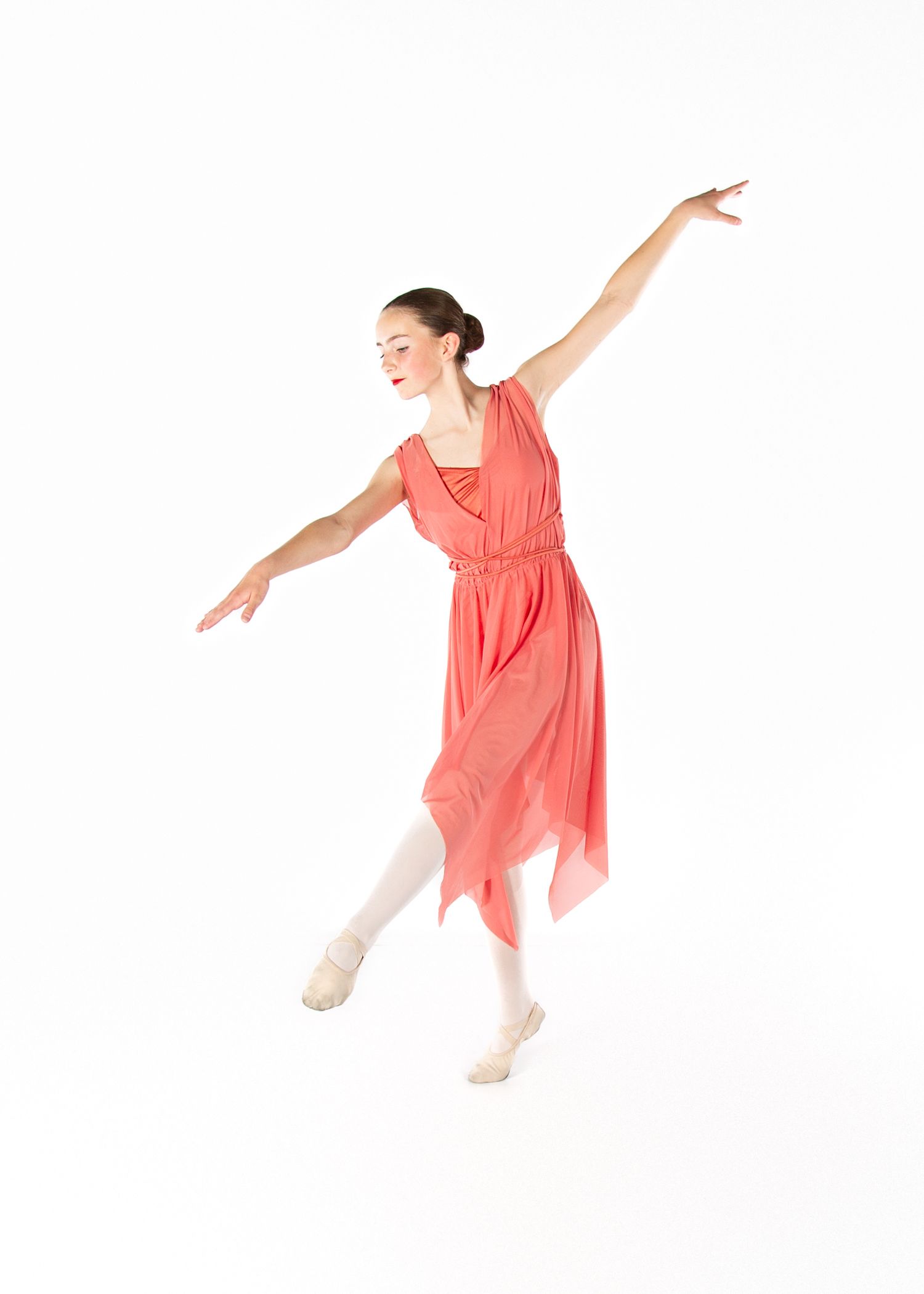 Dancer in flowing coral dress and white tights performs graceful ballet pose with outstretched arms against white backdrop.