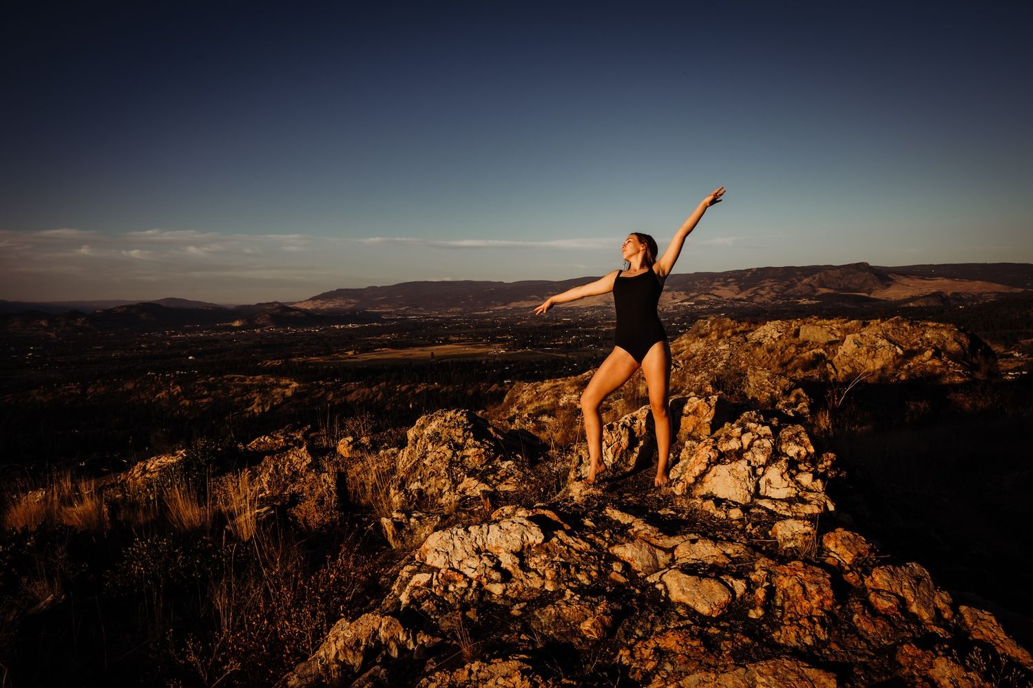Silhouetted dancer poses gracefully atop rocky mountain overlook during golden hour sunset.
