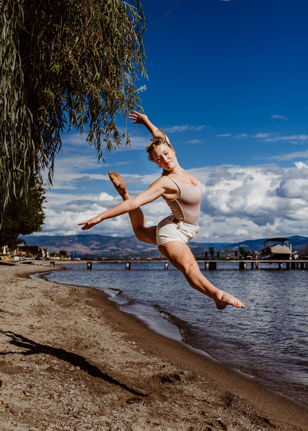A dancer performs an elegant leap at the beach with mountains and a dock in the background on a sunny day.