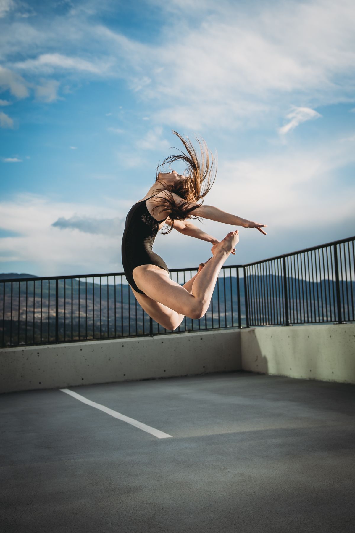 Dancer in black leotard performs graceful leap on rooftop parking area against blue sky and mountain backdrop.