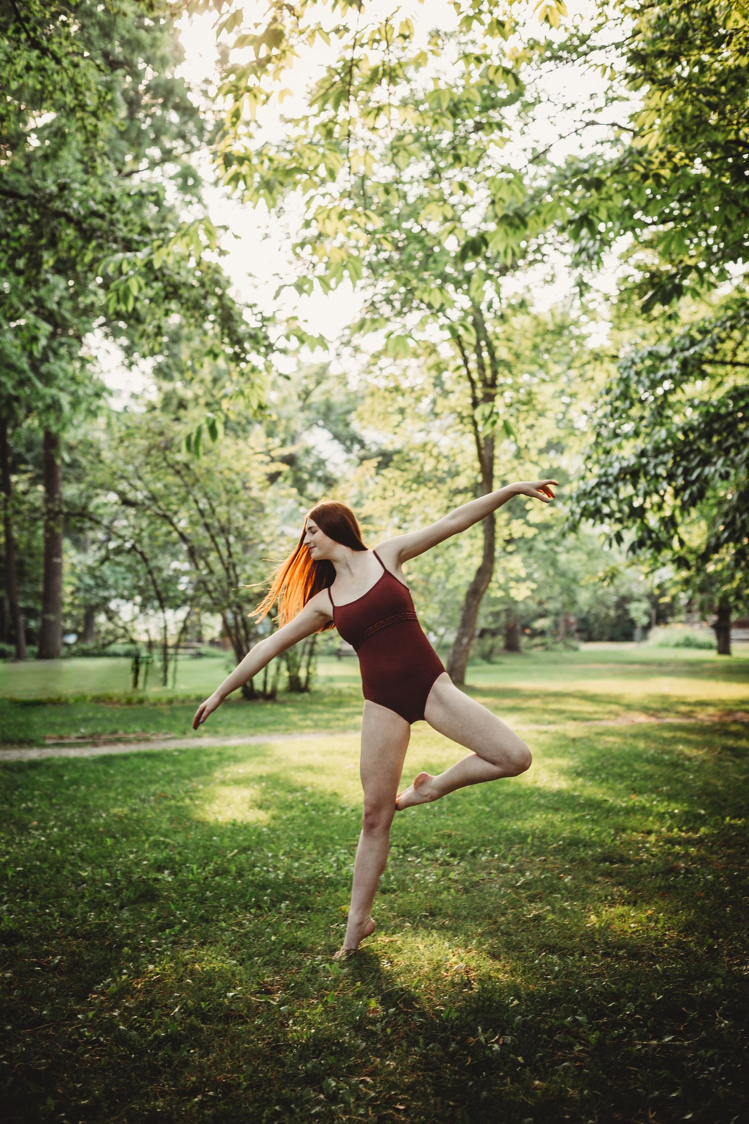 A dancer in a burgundy leotard performs a graceful pose on one leg in a sunlit garden setting.