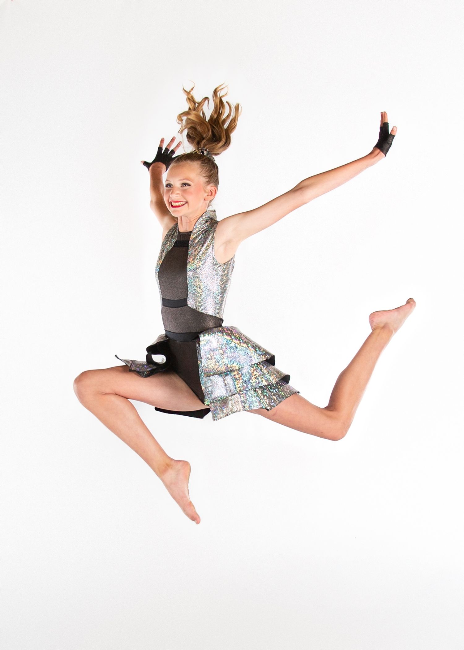 Dancer in sparkling gray costume performs energetic leap with hair flying and arms outstretched against white background.