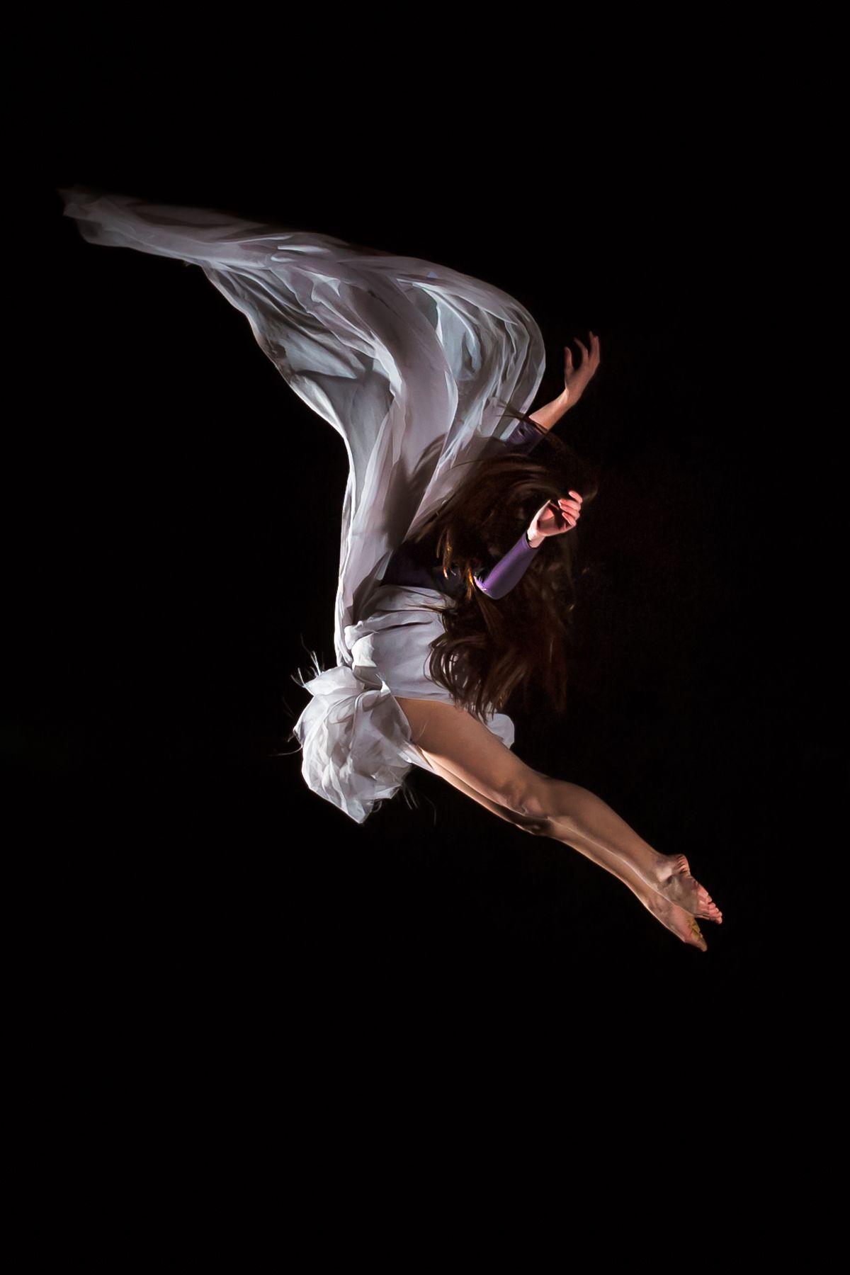 Graceful dancer in white flowing fabric leaps through the air against a dark background.