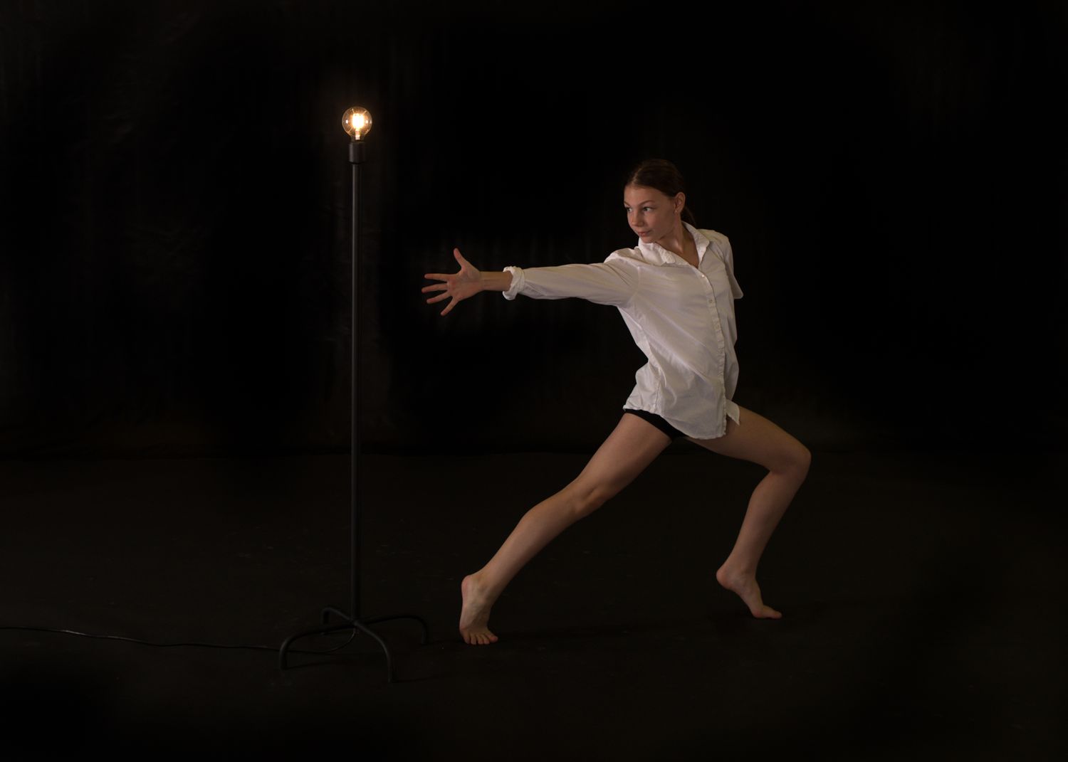 Dancer in white shirt performs dramatic leap against dark background with single illuminated streetlight.