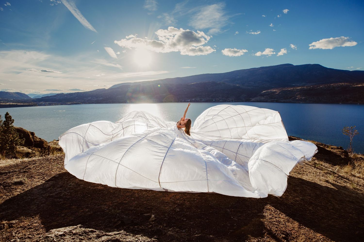 White blanket and pillow setup on cliff overlooking scenic lake with sunlight reflecting off water and mountains.