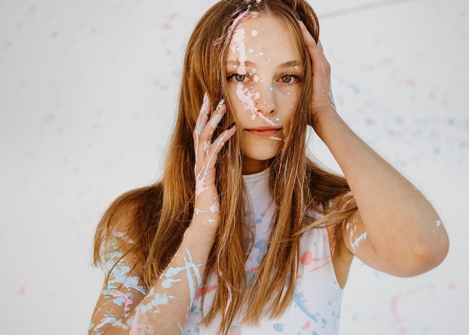 Person with long brown hair and freckles wearing a white top poses with hand against head against white wall.