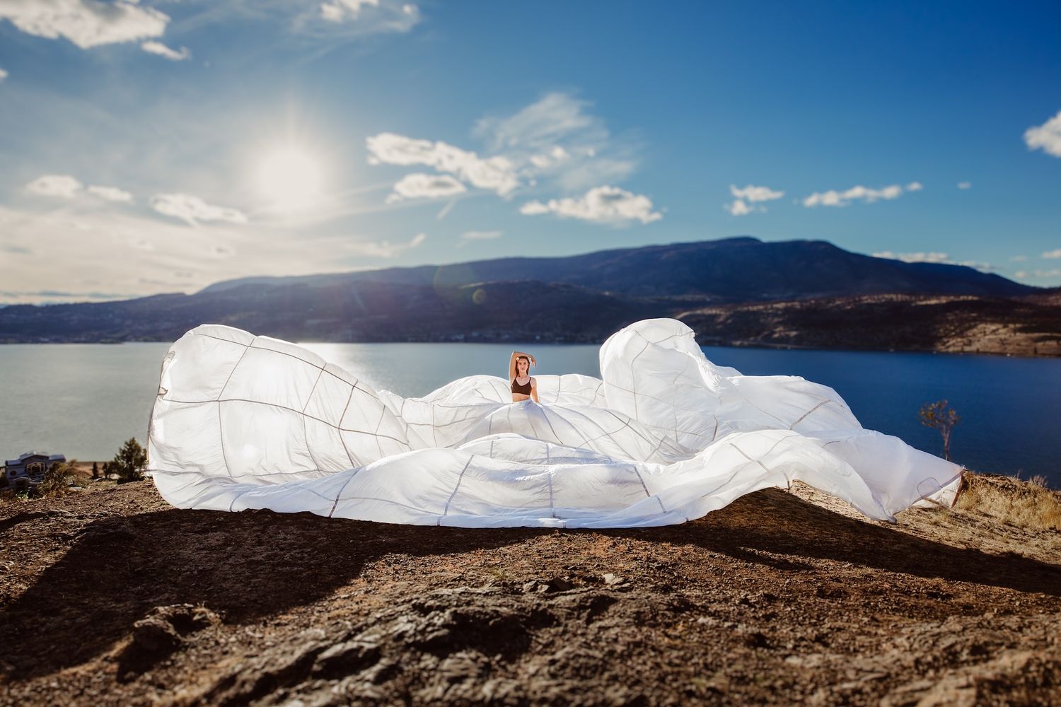 Giant white inflatable art installation rests on rocky overlook against scenic mountain lake backdrop under blue skies.