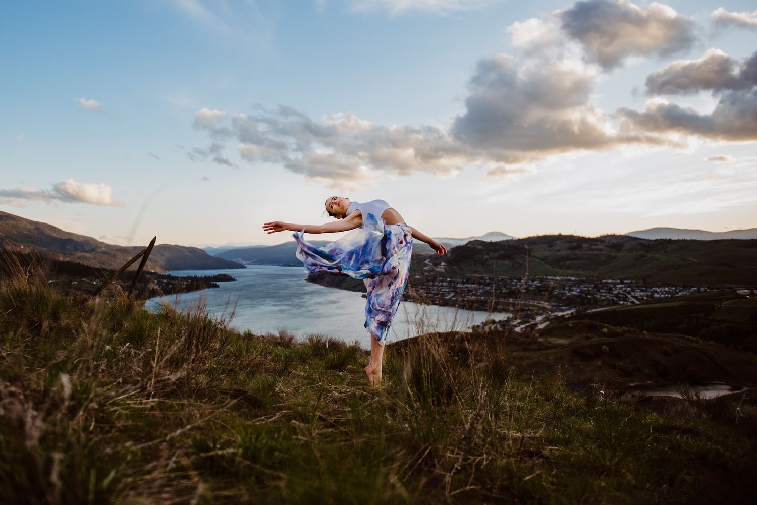 A person performs a martial arts kick pose on a hilltop overlooking a scenic coastal landscape at sunset.