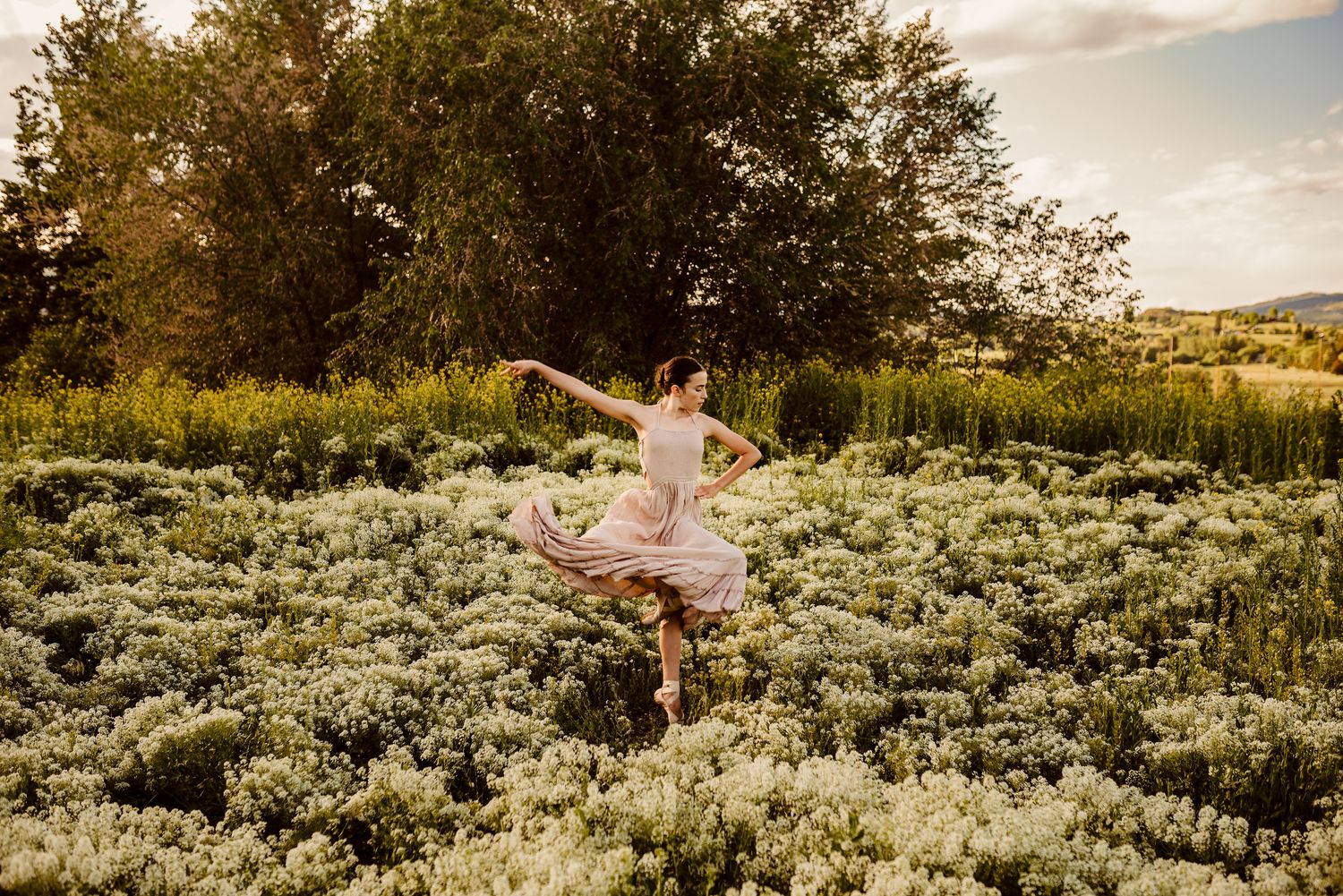 A graceful dancer twirls in a pink dress among a field of white wildflowers at golden hour.