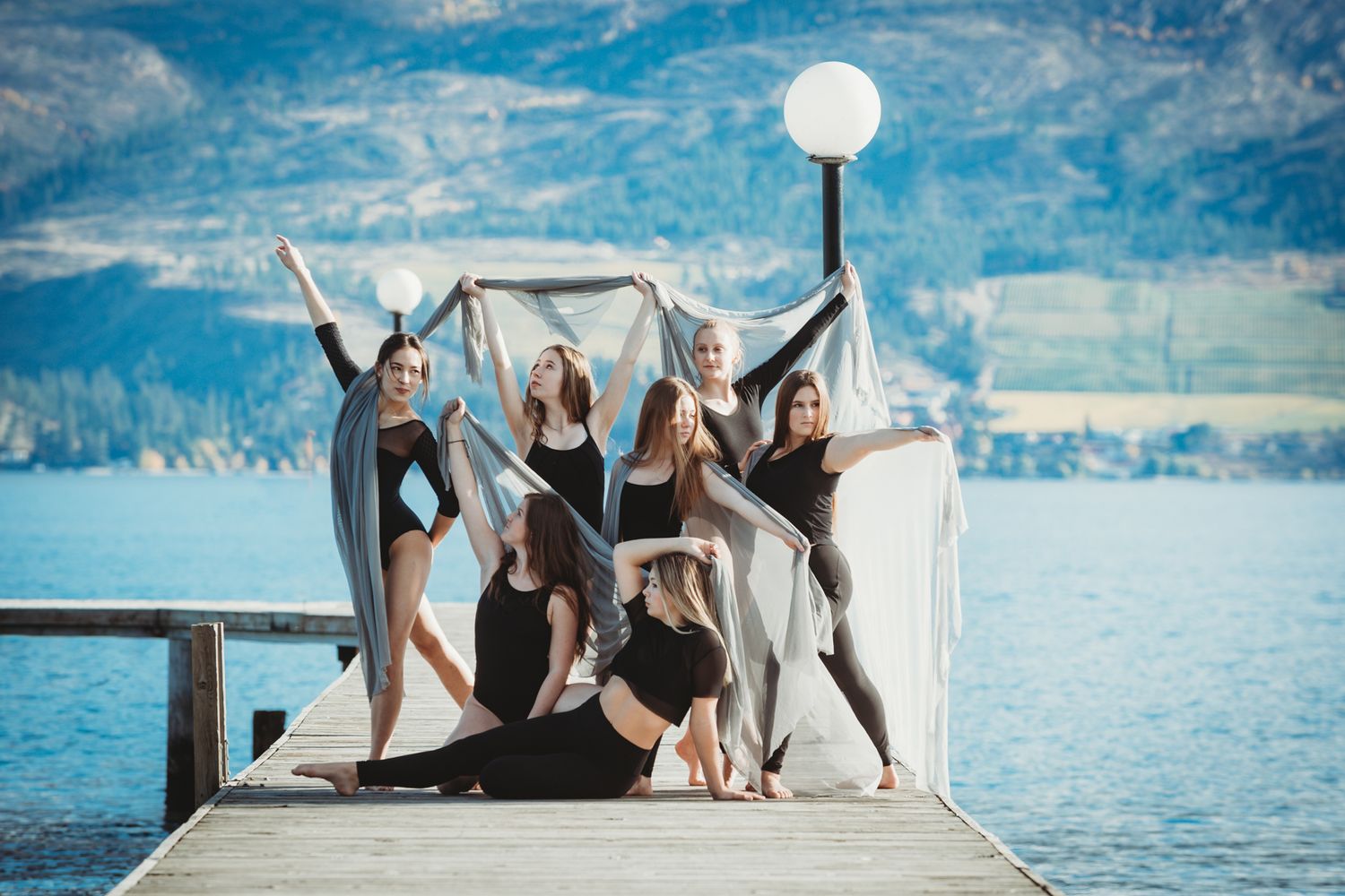 A group of dancers in black leotards pose gracefully on a dock with a scenic mountain lake backdrop.